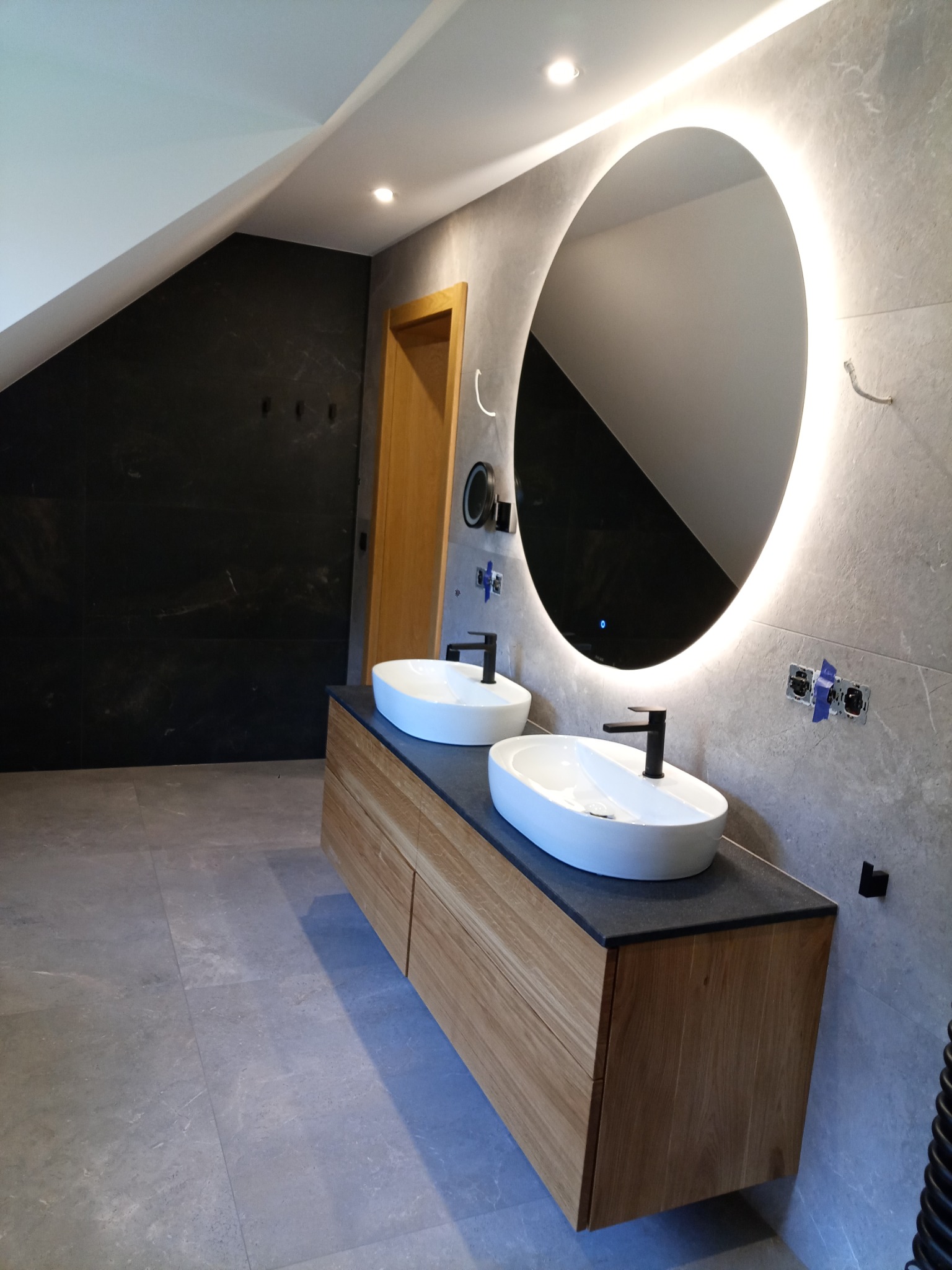 Nowoczesna łazienka na poddaszu z owalnym lustrem LED, dwiema umywalkami nablatowymi na drewnianej szafce i szarą płytką imitującą kamień na ścianach i podłodze.