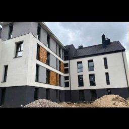 USŁUGI REMONTOWO-BUDOWLANE Mariusz Łatka - Nowoczesny budynek po ociepleniu elewacji w odcieniach bieli i szarości z drewnianymi panelami. Na pierwszym planie hałda piasku i kamieni.