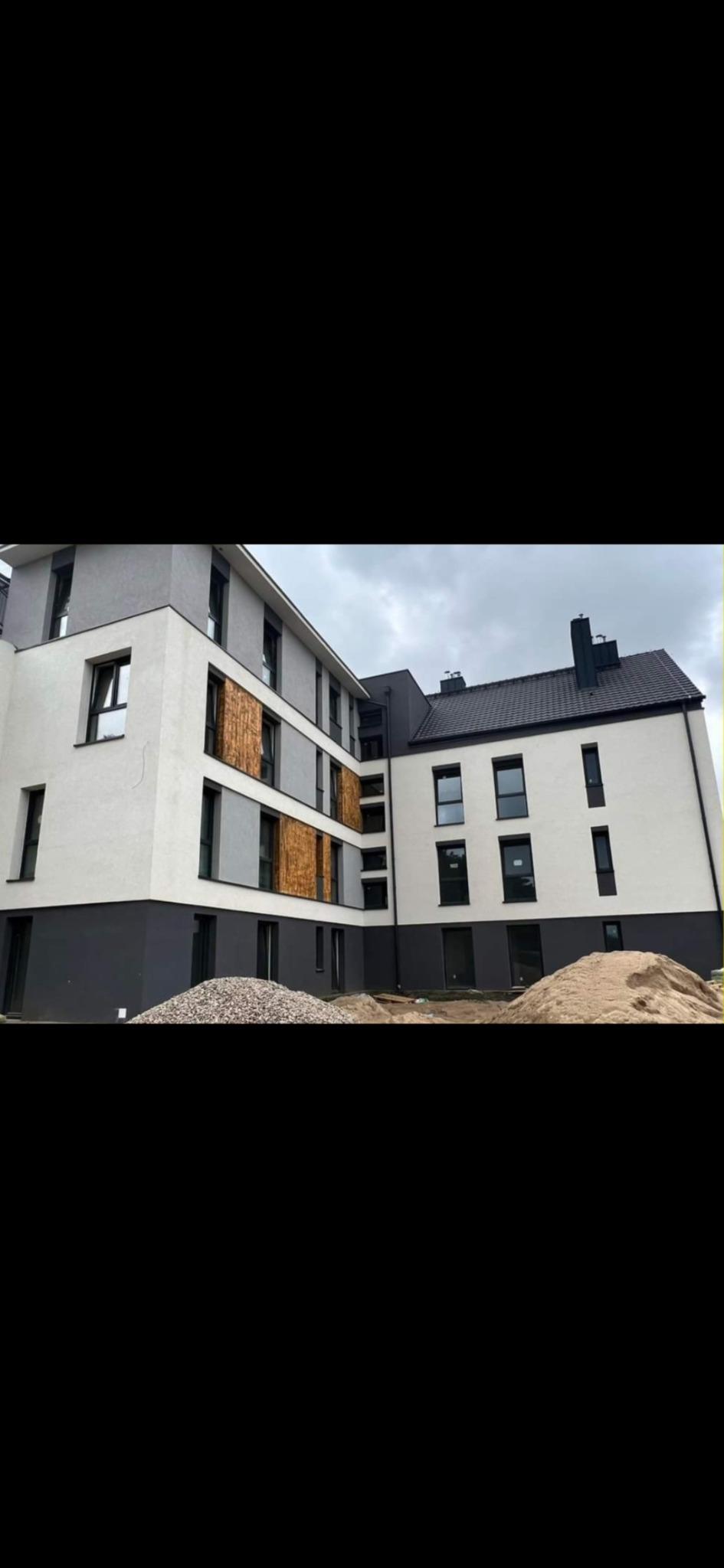 Nowoczesny budynek po ociepleniu elewacji w odcieniach bieli i szarości z drewnianymi panelami. Na pierwszym planie hałda piasku i kamieni.