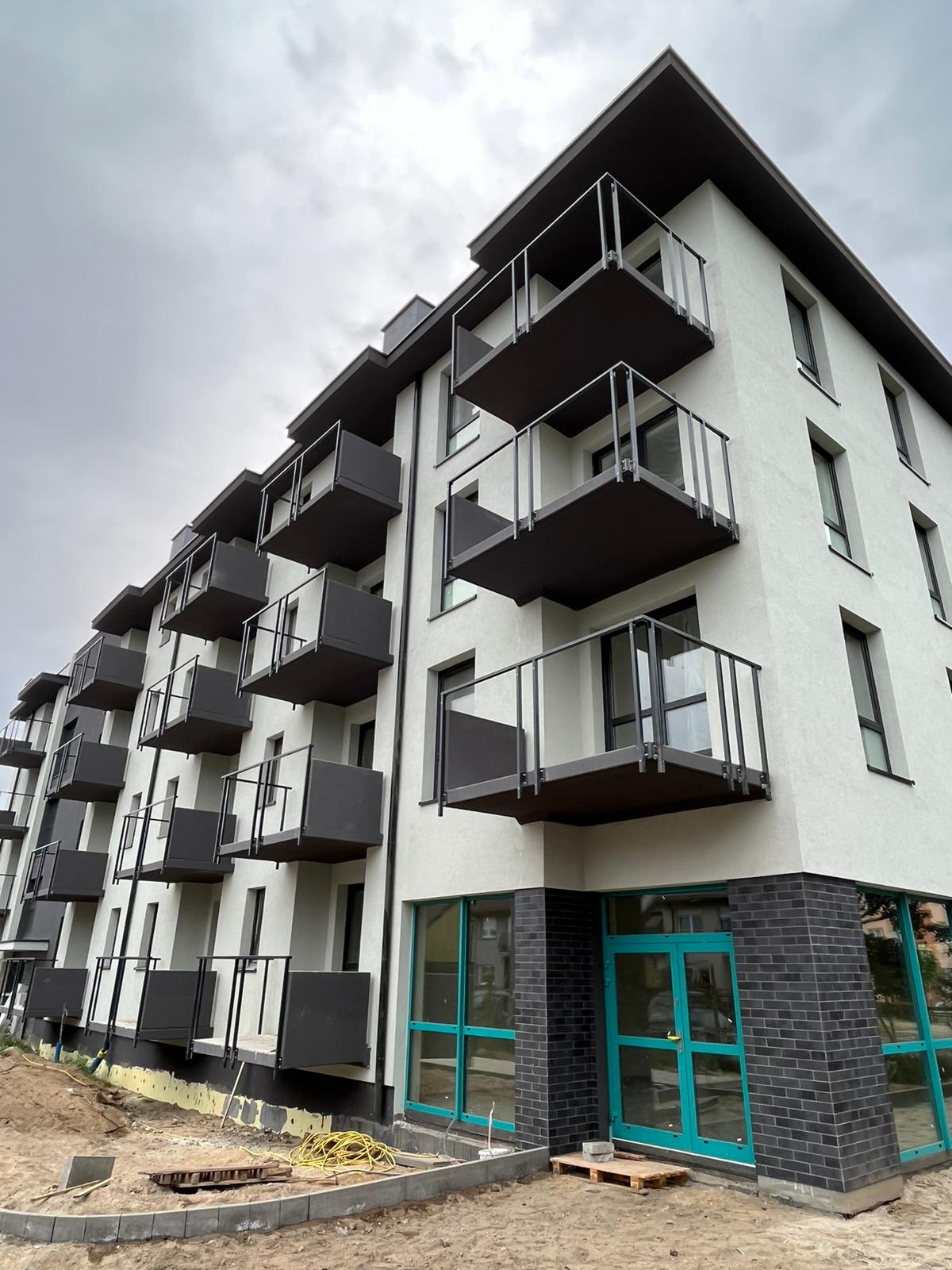 Nowoczesny blok z balkonami w Maszkowie. Biała elewacja i czarne balustrady balkonów. Widoczne prace wykończeniowe wokół budynku.