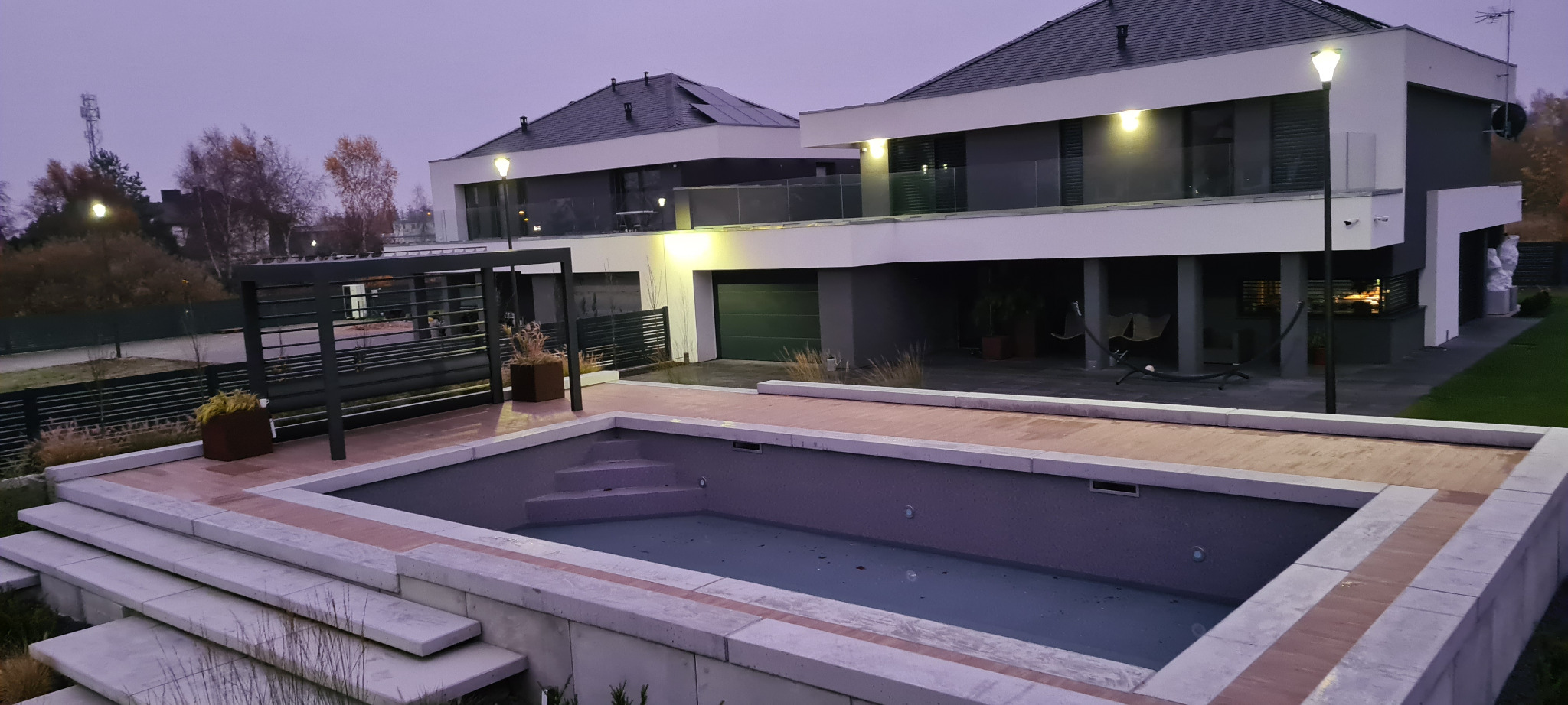 Nowoczesny dom z basenem w szaro-białej tonacji. Basen z jasnymi schodami i drewnianym obramowaniem. Oświetlenie zewnętrzne dodaje ciepła. Widok na budynek z garażem.