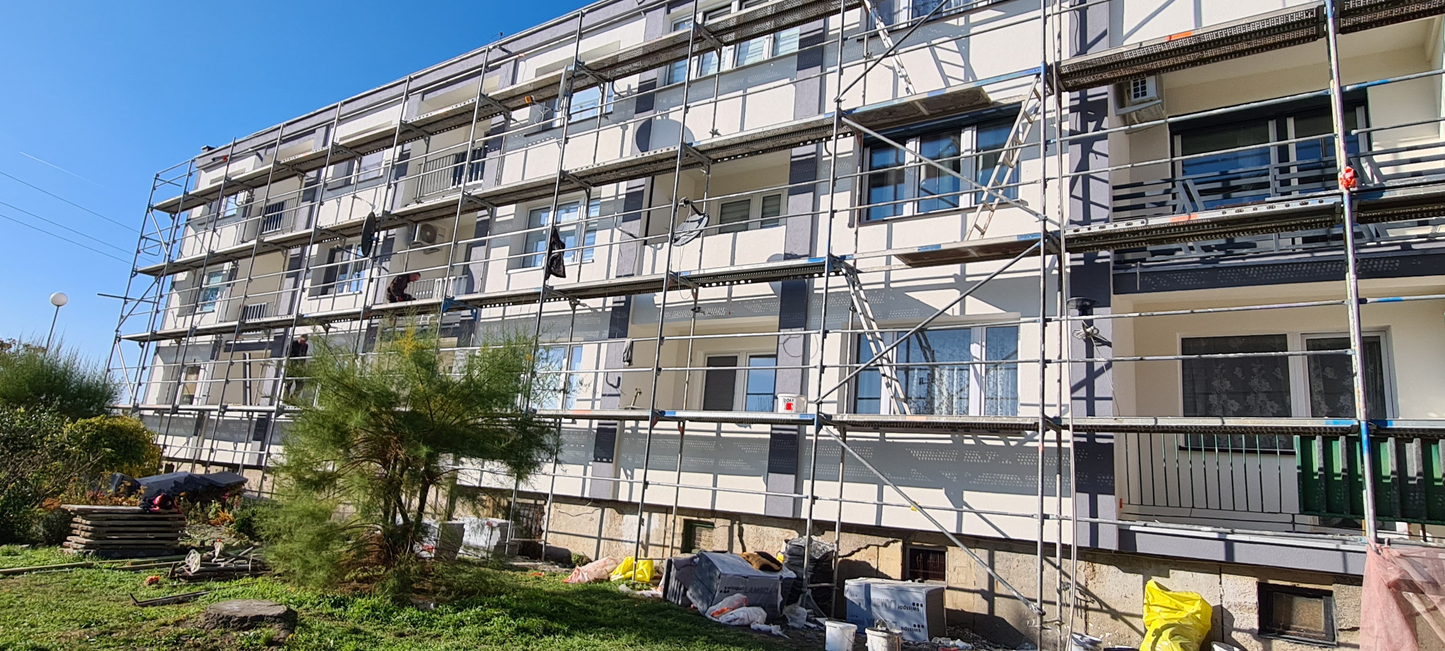 Wielopiętrowy budynek w rusztowaniach, elewacja w trakcie odnawiania. Widoczne materiały budowlane, drzewa i fragment trawnika. Słoneczny dzień.