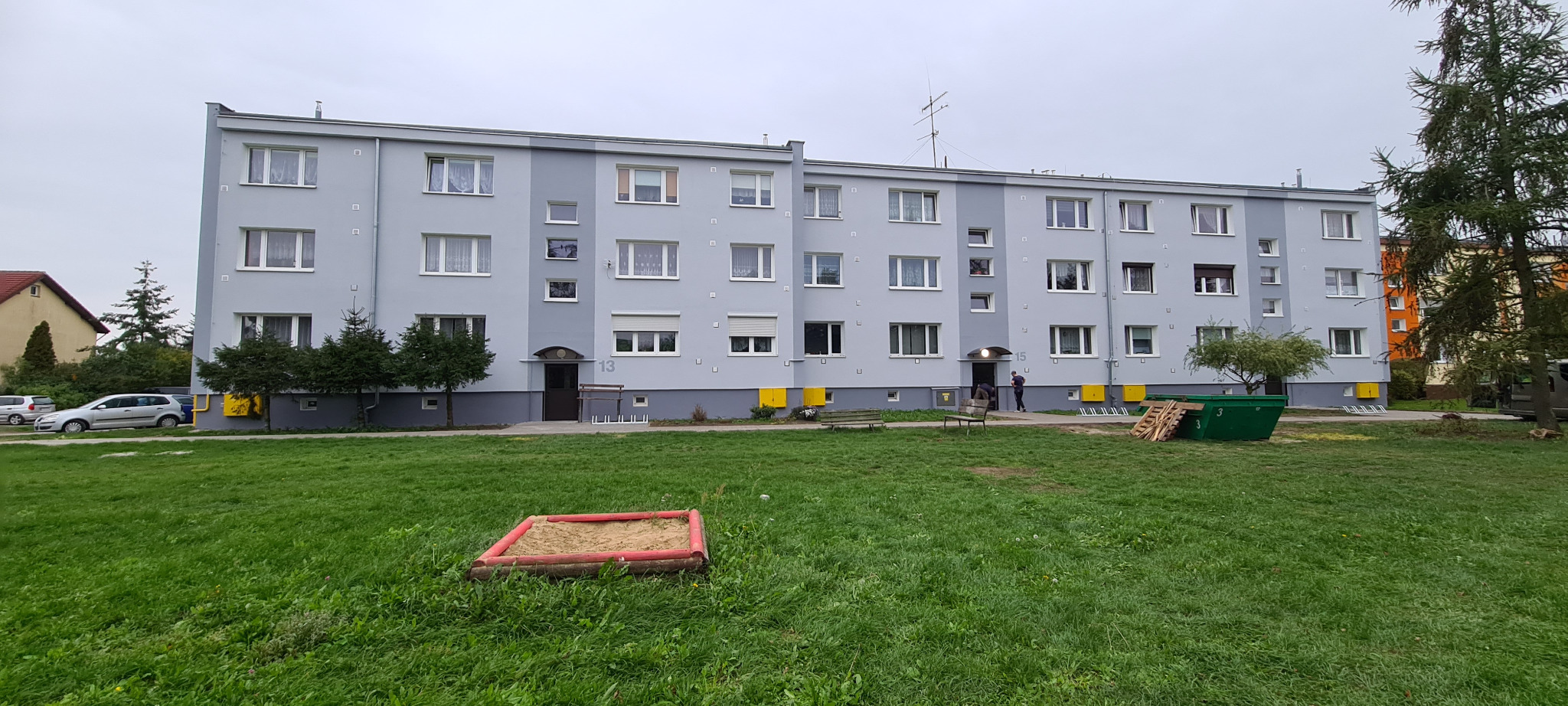 Wielorodzinny budynek po odświeżeniu elewacji w odcieniach szarości, z numerami 13 i 15 nad wejściami. Na pierwszym planie trawnik i piaskownica.