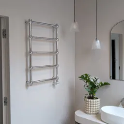 Nowoczesna łazienka z grzejnikiem drabinkowym, umywalką nablatową i podłogą w odcieniach beżu. Dwie lampy wiszące i roślina doniczkowa dodają przytulności.