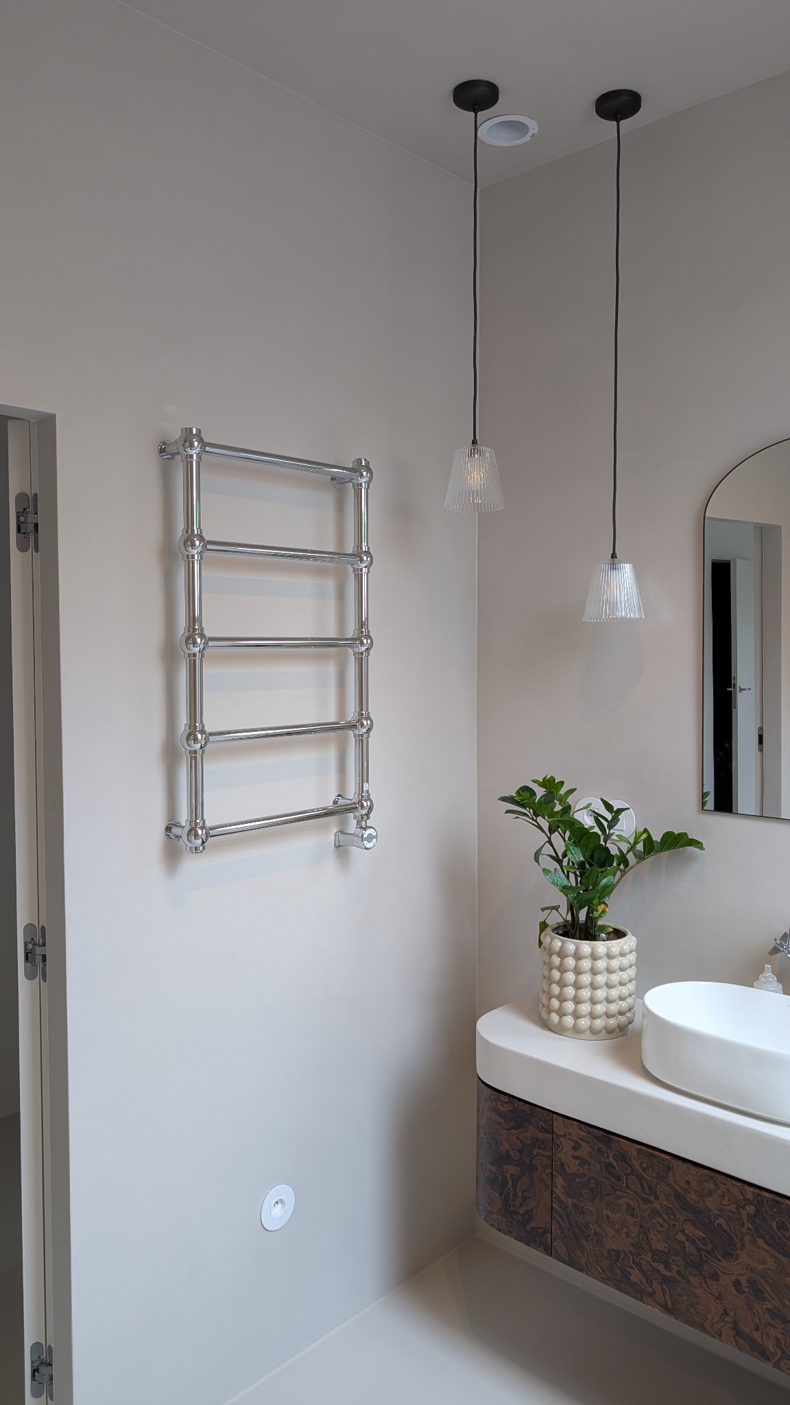 Nowoczesna łazienka z grzejnikiem drabinkowym, umywalką nablatową i podłogą w odcieniach beżu. Dwie lampy wiszące i roślina doniczkowa dodają przytulności.