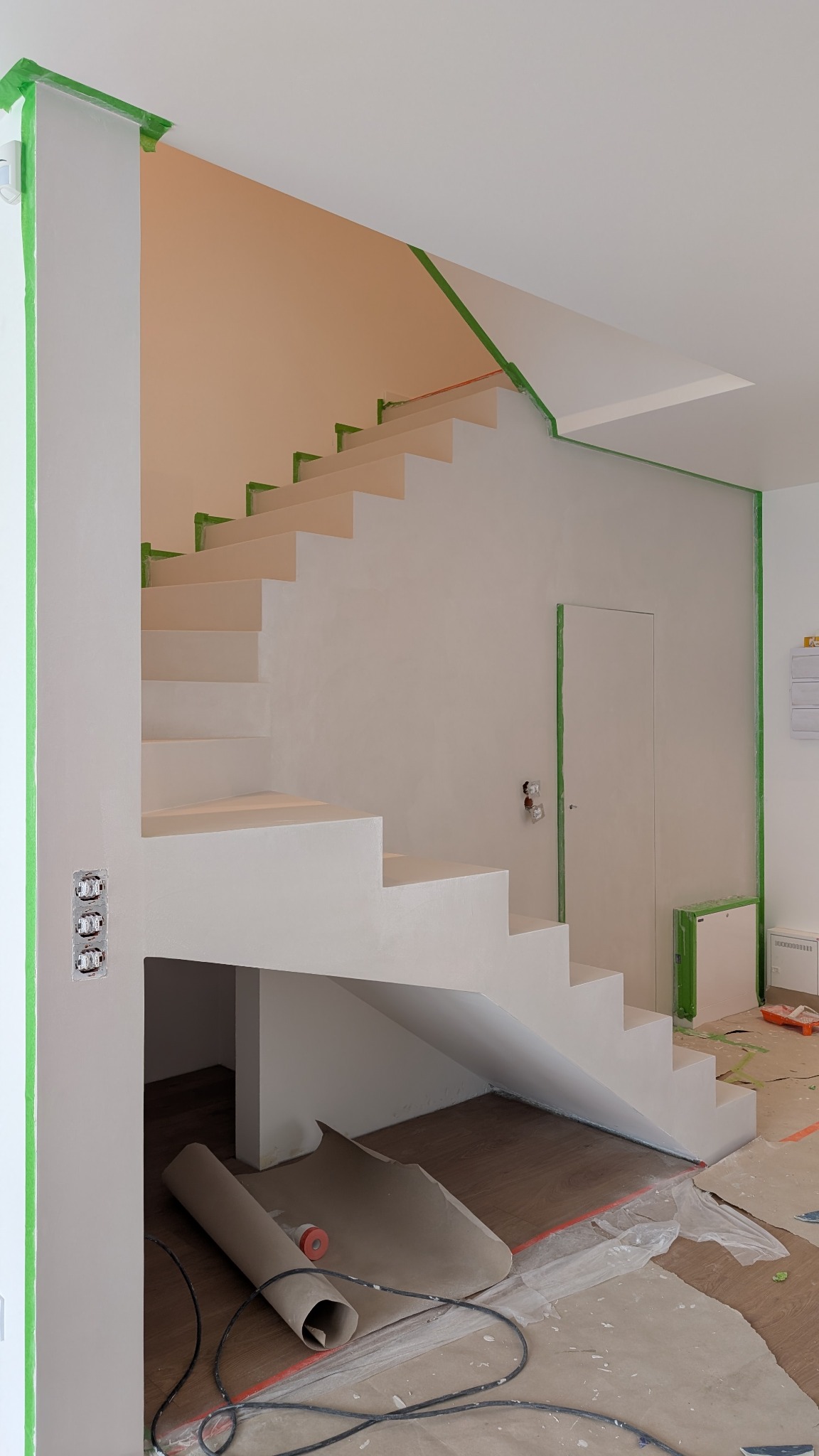 Wykończone schody w jasnym odcieniu, zabezpieczone taśmą malarską, z widocznym fragmentem podłogi i narzędziami malarskimi, ukazujące etap prac wykończeniowych.