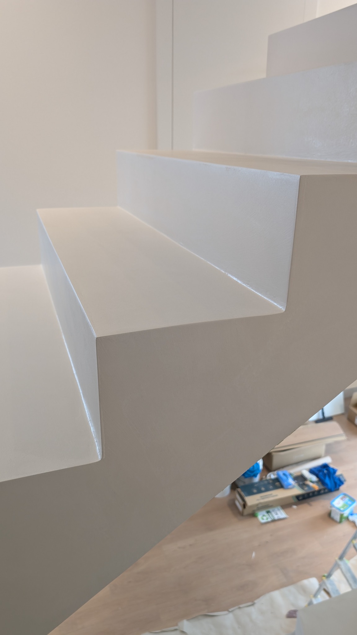 Białe schody z wykończeniem z gładzi, widoczne z bliska, z narzędziami malarskimi w tle. Minimalistyczny design, jasne wnętrze, subtelne oświetlenie.