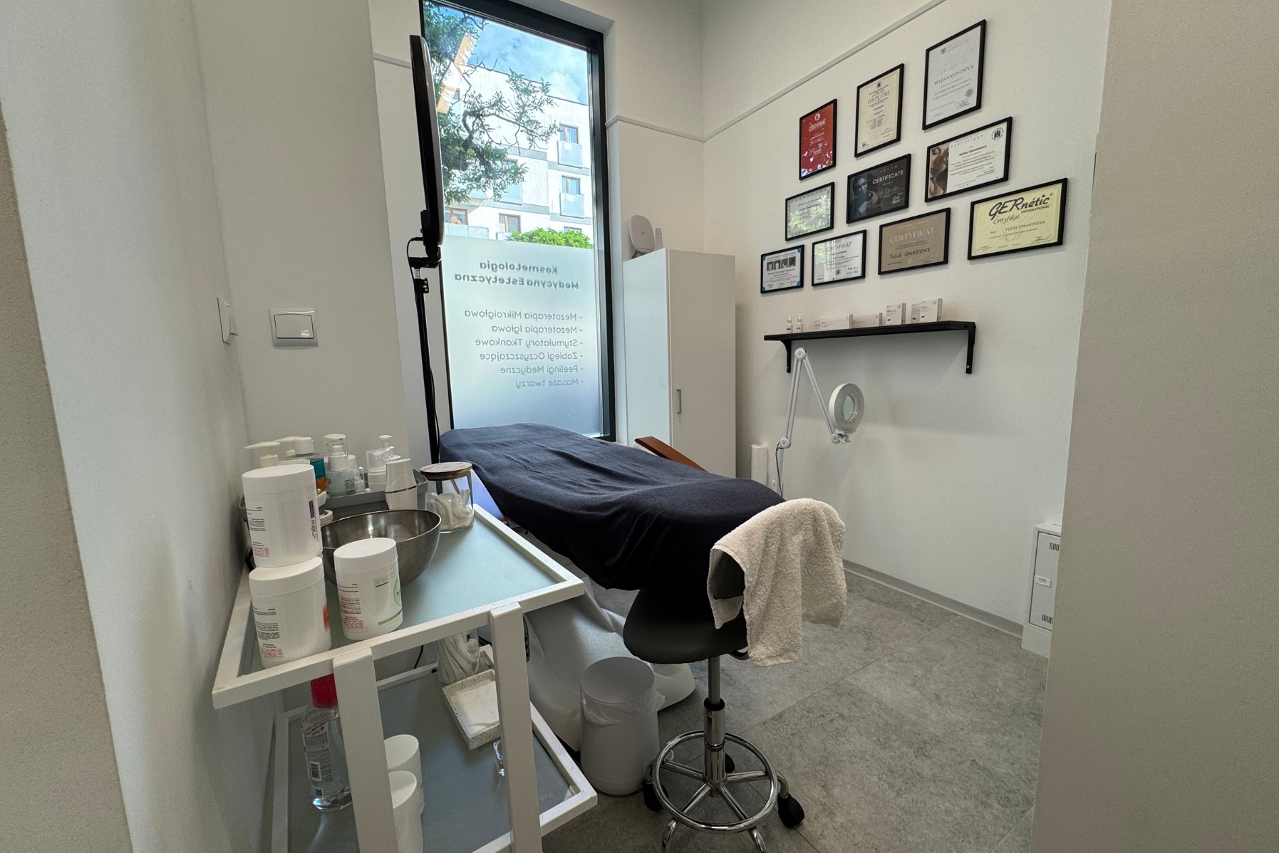 Jasne wnętrze gabinetu: leżanka kosmetyczna z ciemnym nakryciem, stolik z akcesoriami, krzesło i ściana z certyfikatami. Widok na zewnątrz przez okno.