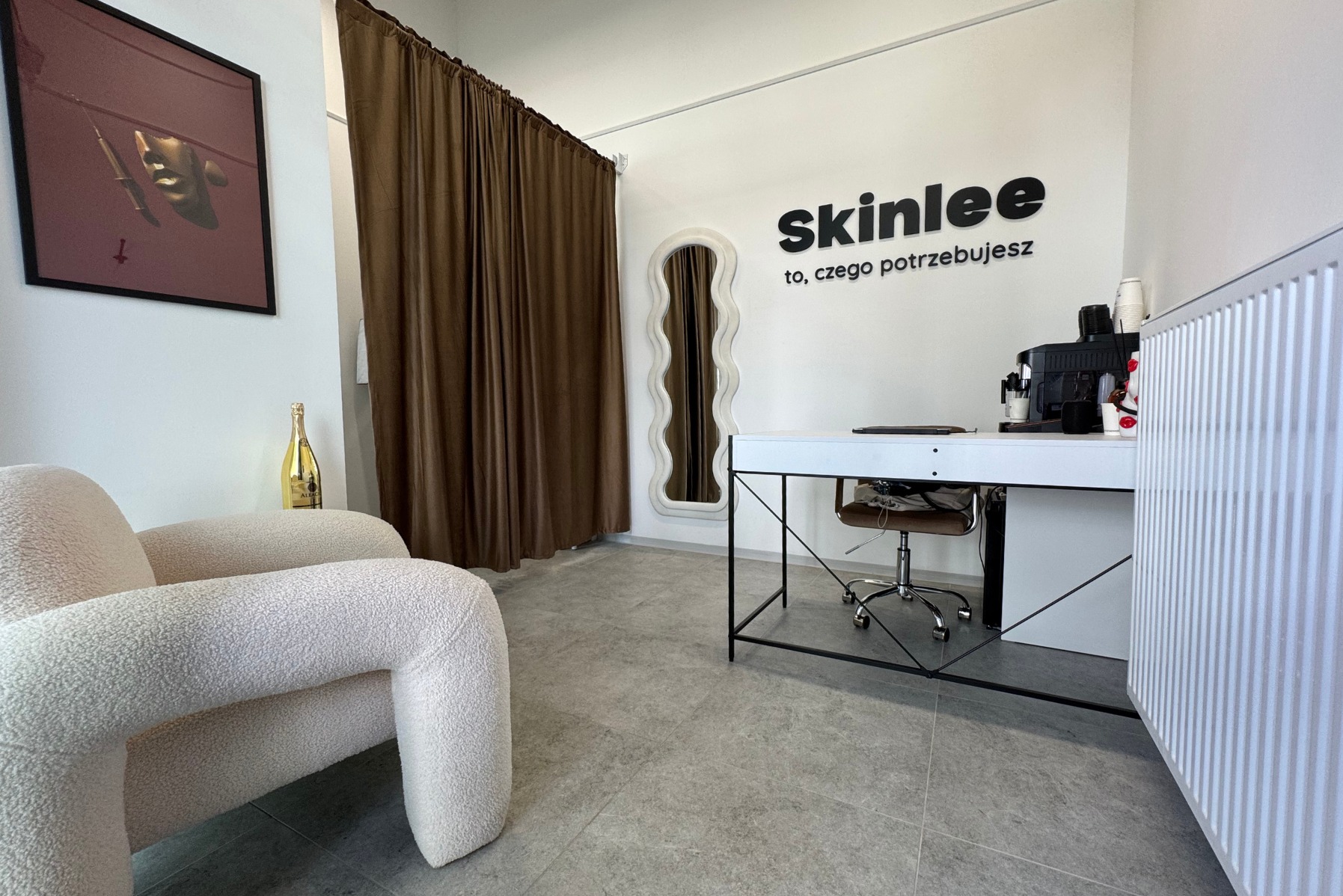 Wnętrze salonu kosmetycznego Skinlee: fotel boucle, lustro o falistym kształcie, biurko z ekspresem do kawy i logo na ścianie. Nowoczesny design i przytulna atmosfera.