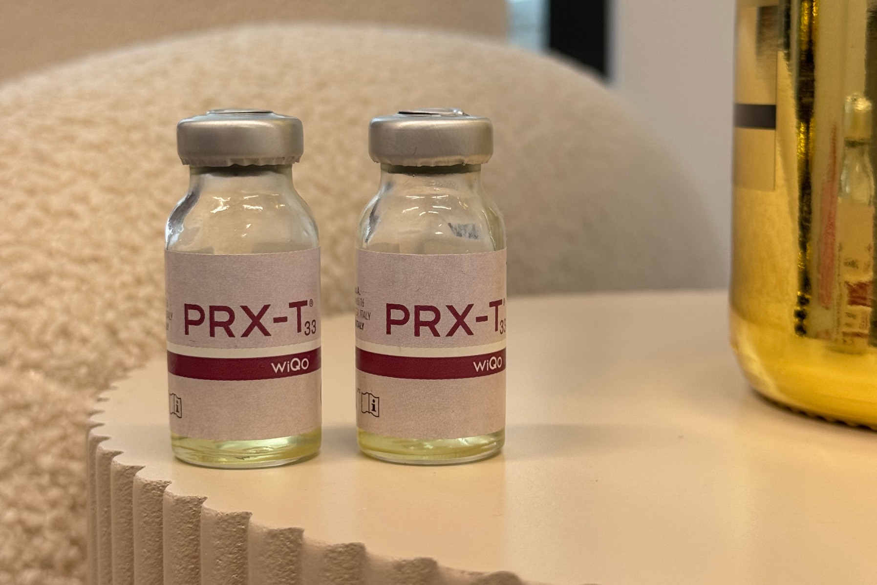 Dwie fiolki PRX-T33 na stoliku o falistej strukturze, z rozmytym tłem w odcieniach beżu i fragmentem złotego elementu dekoracyjnego po prawej stronie.