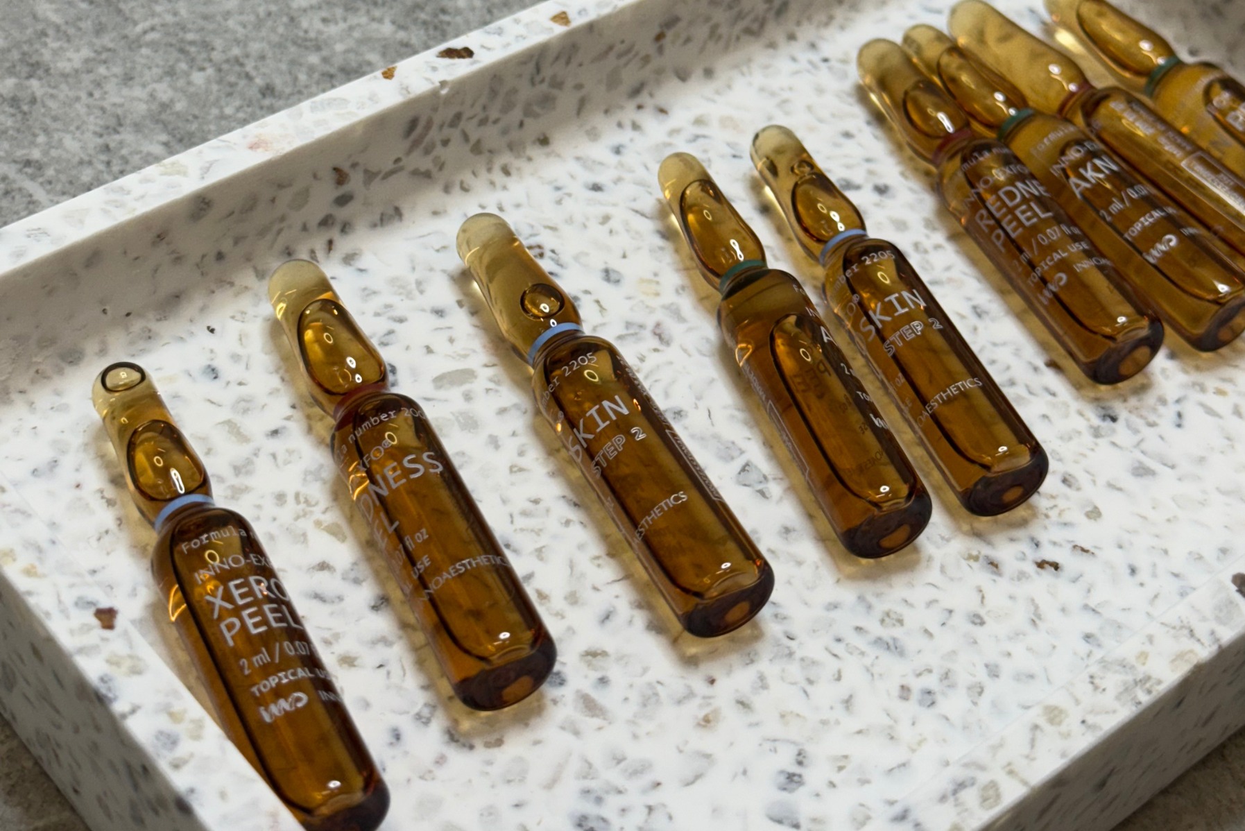 Szklane ampułki z preparatami do peelingu chemicznego na marmurowej tacy. Widoczne napisy 'Xero Peel', 'Skin Step 2' i 'Redne Peel'.