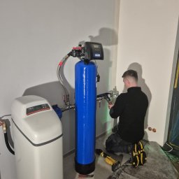 AQUA-MAT - Instalacja uzdatniacza wody: hydraulik w czarnym ubraniu łączy rury do niebieskiego filtra, obok biały zbiornik na sól. Na podłodze narzędzia i mata ochronna.