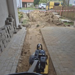 AQUA-MAT - Układanie kostki brukowej: zagęszczarka w rowie, przygotowanie podłoża. Częściowo ułożona kostka, hałda piasku, ogrodzenie z taśmą ostrzegawczą w tle.