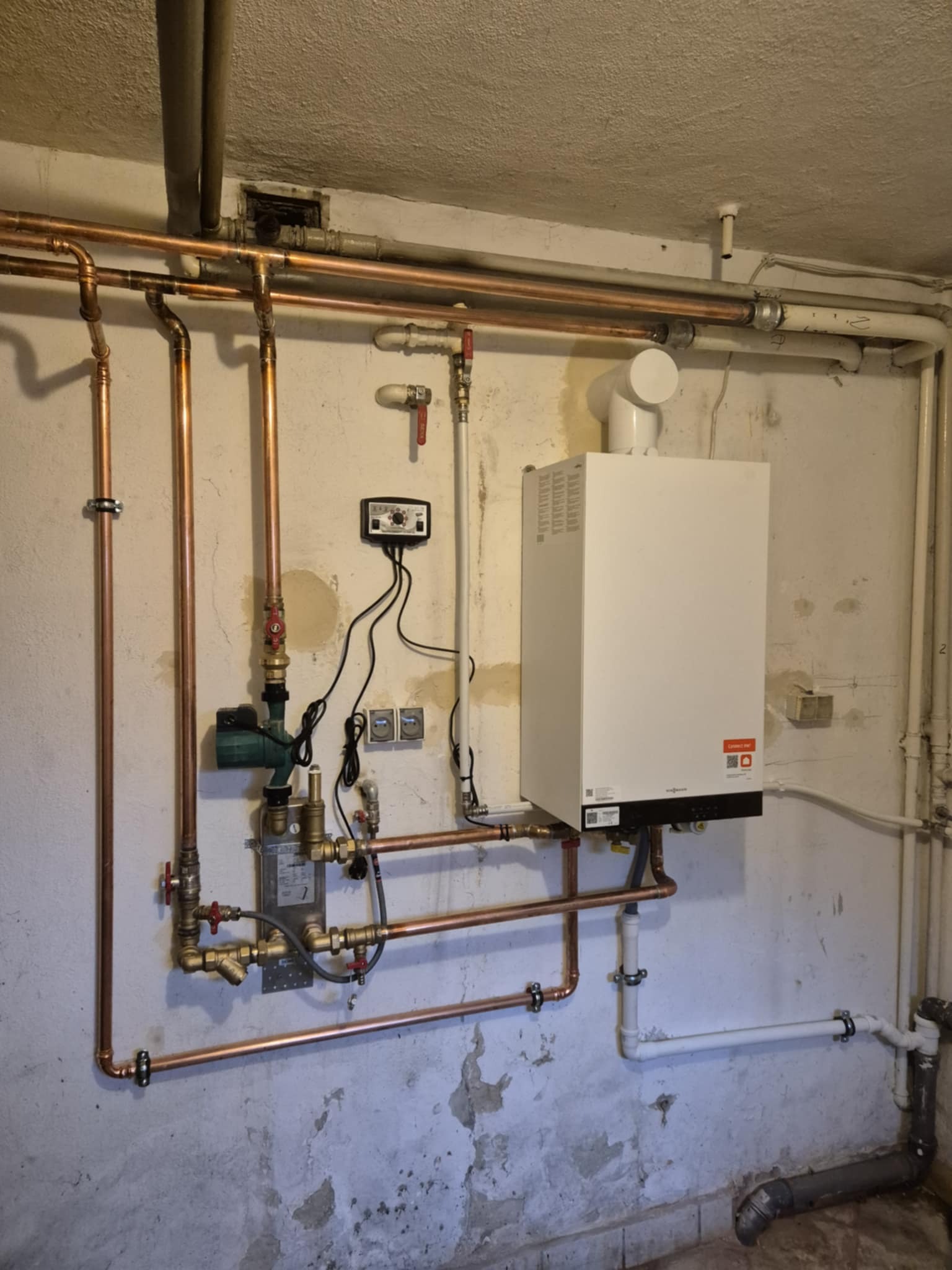 Biały piec CO na ścianie z instalacją z miedzianych rur, zaworami i pompą. Widoczne przewody elektryczne i regulator. Tło stanowi ściana z widoczną strukturą.