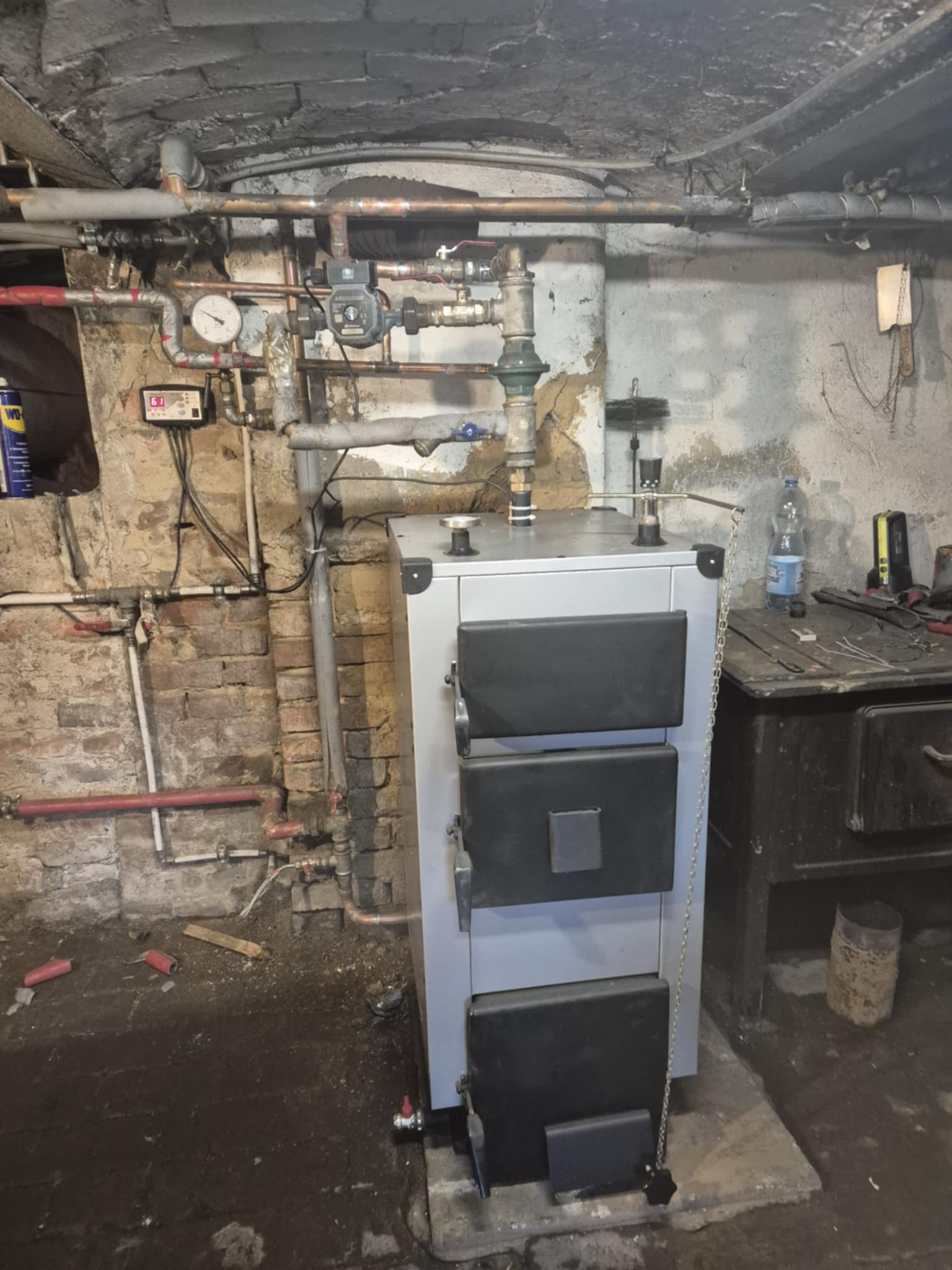 Stary piec CO z instalacją hydrauliczną w piwnicy, widoczne rury, zawory, manometr i regulator. Na stole narzędzia i butelka wody. Nierówna ceglana ściana.