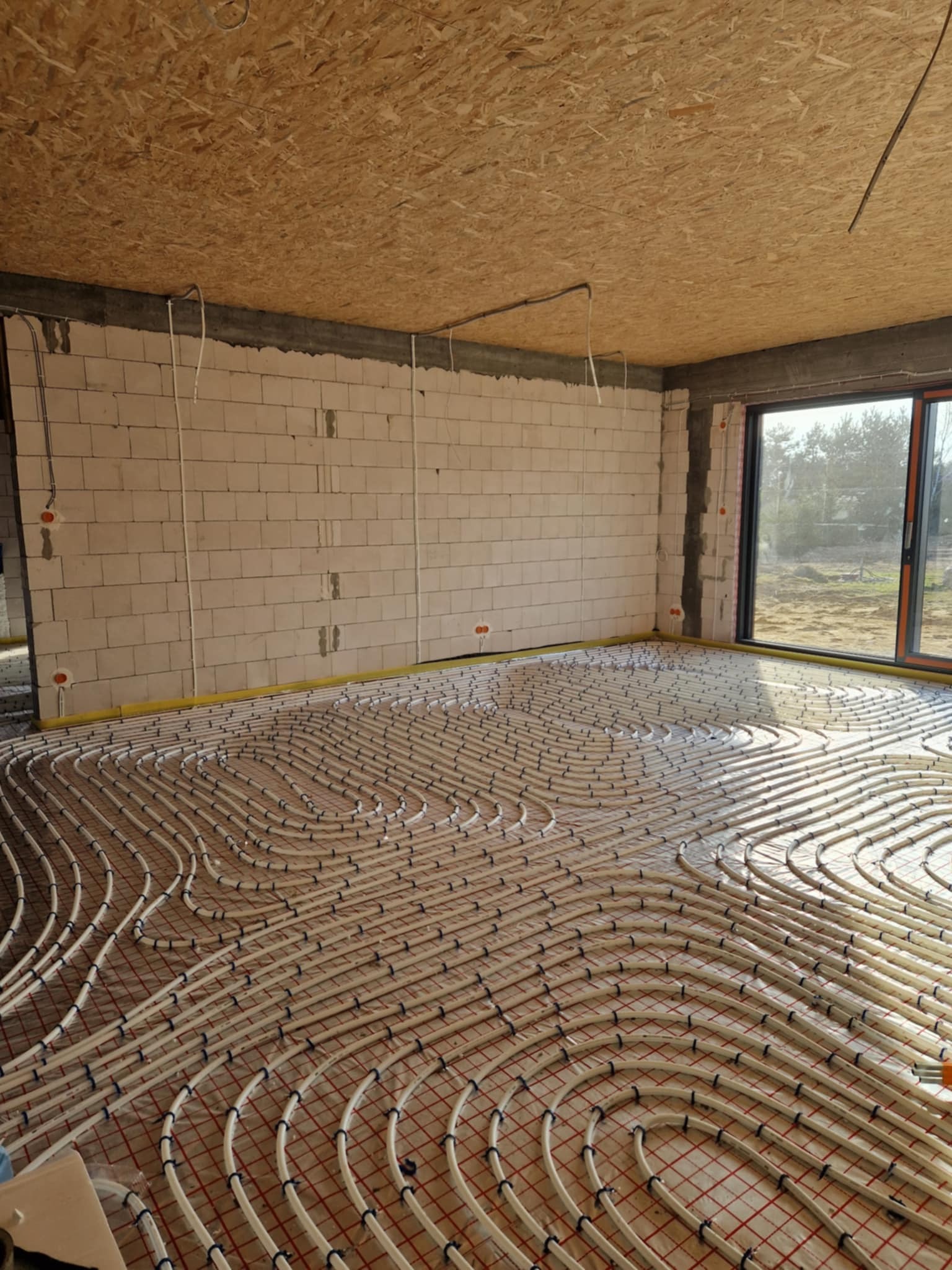 Białe rury ogrzewania podłogowego ułożone na czerwonej siatce w surowym wnętrzu z oknem i ścianami z białych bloczków. Widoczny sufit z płyty OSB.