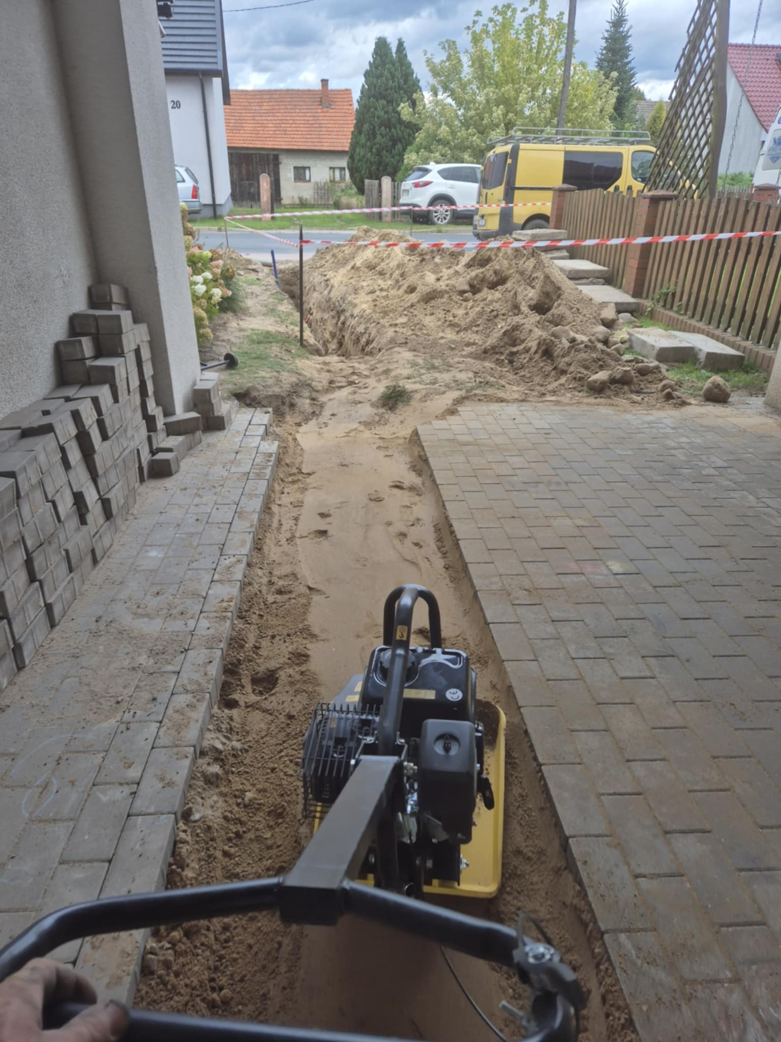 Układanie kostki brukowej: zagęszczarka w rowie, przygotowanie podłoża. Częściowo ułożona kostka, hałda piasku, ogrodzenie z taśmą ostrzegawczą w tle.