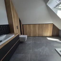 Nowoczesna łazienka na poddaszu z drewnianą szafą w zabudowie, czarną umywalką i szarą podłogą. Okno dachowe wpuszcza jasne światło.