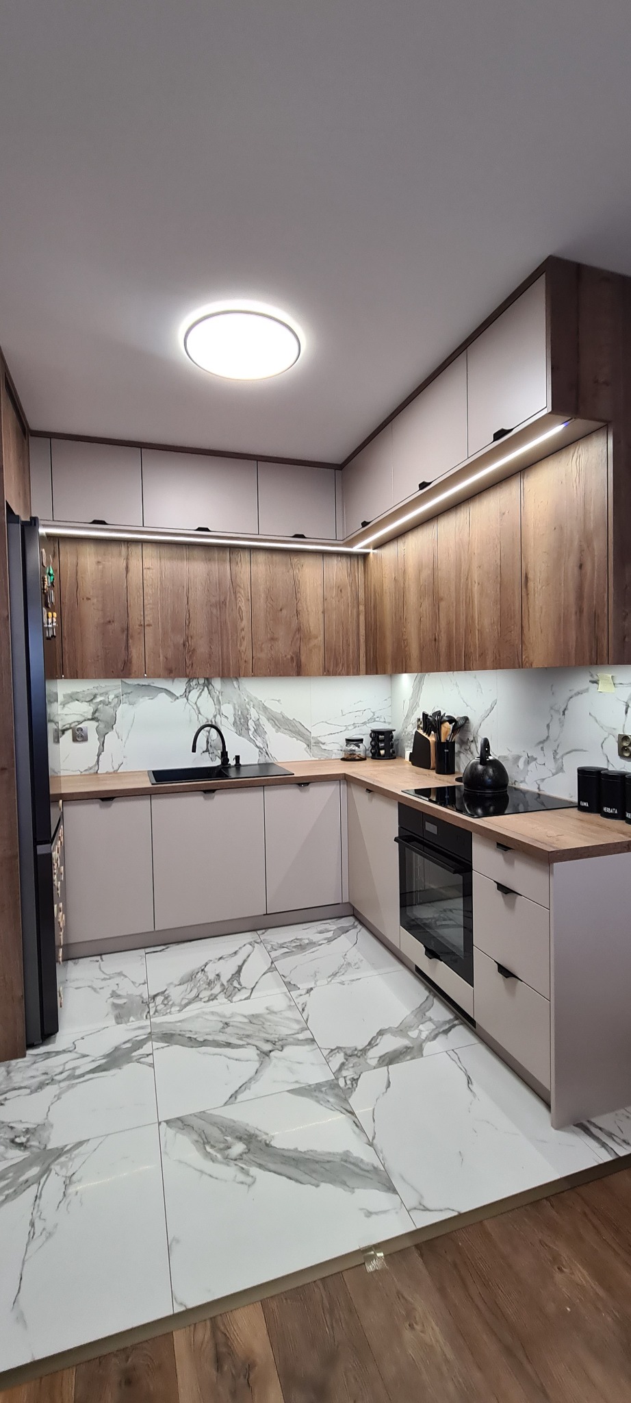 Nowoczesna kuchnia w kształcie litery L z drewnianymi i białymi szafkami, marmurowymi płytkami na podłodze i ścianie. Oświetlenie LED pod szafkami górnymi.