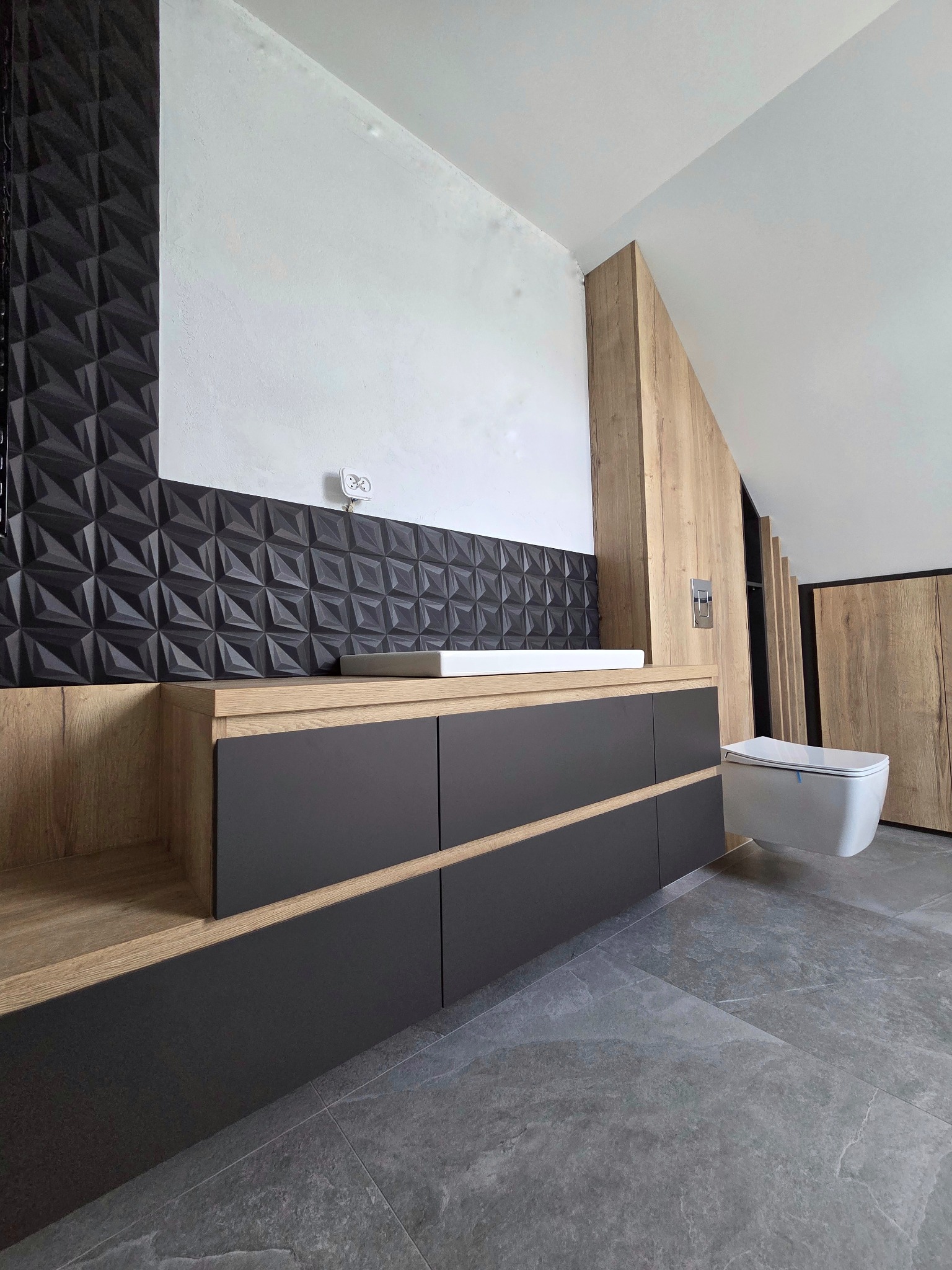 Nowoczesna łazienka na poddaszu: szara zabudowa z umywalką, geometryczna mozaika i podwieszana toaleta. Drewniane akcenty ocieplają wnętrze.