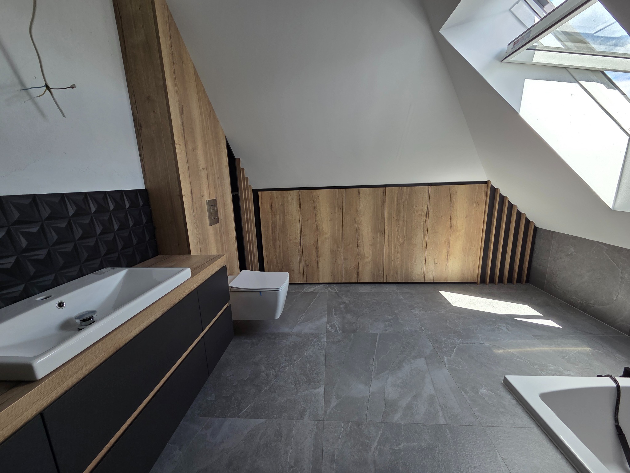 Nowoczesna łazienka na poddaszu z drewnianą szafą w zabudowie, czarną umywalką i szarą podłogą. Okno dachowe wpuszcza jasne światło.