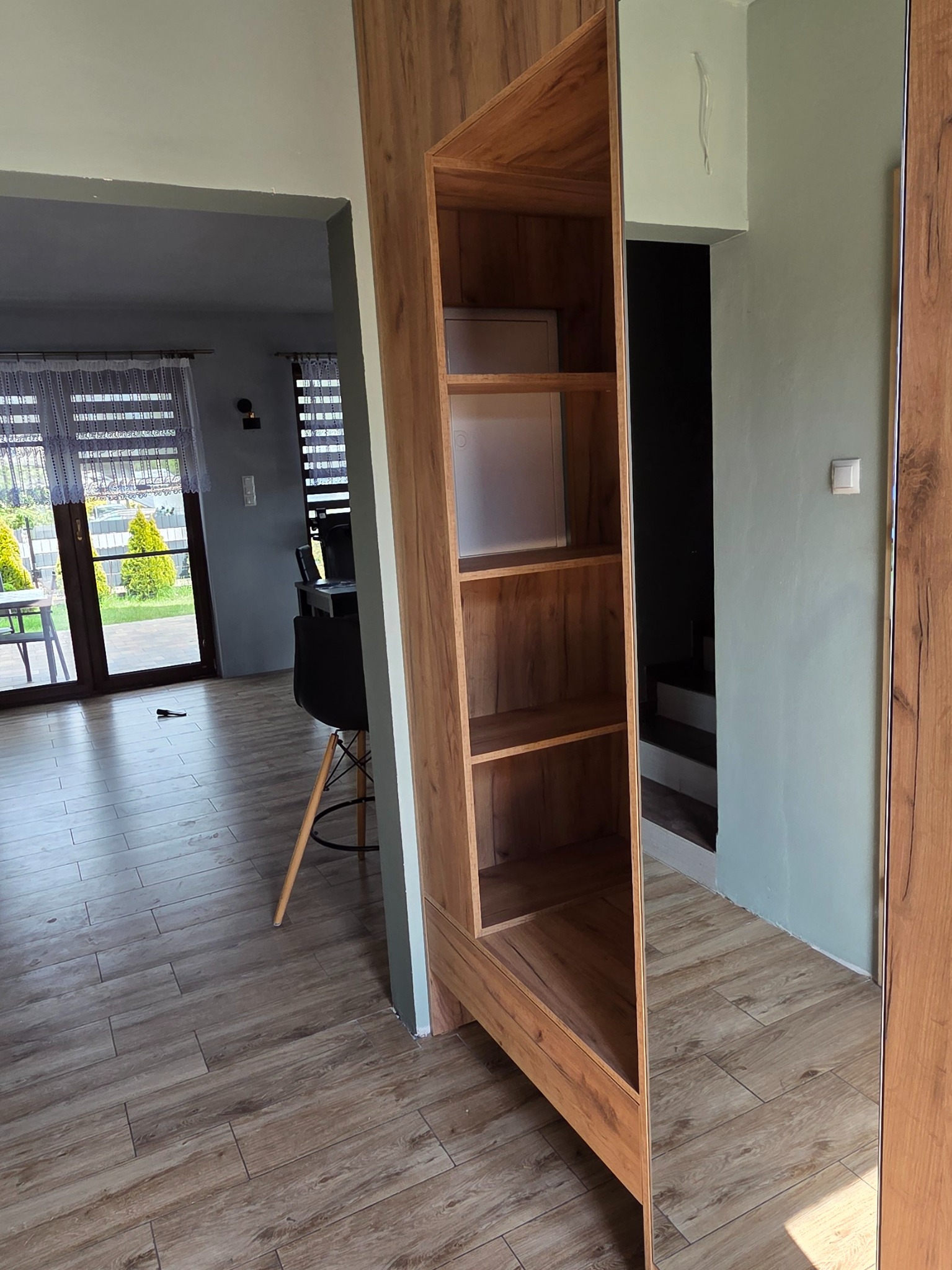 Wysoka szafa wnękowa z lustrem i drewnianymi półkami w jasnym wnętrzu. Widoczny fragment podłogi i okna. Detal wykończenia i designu mebla.