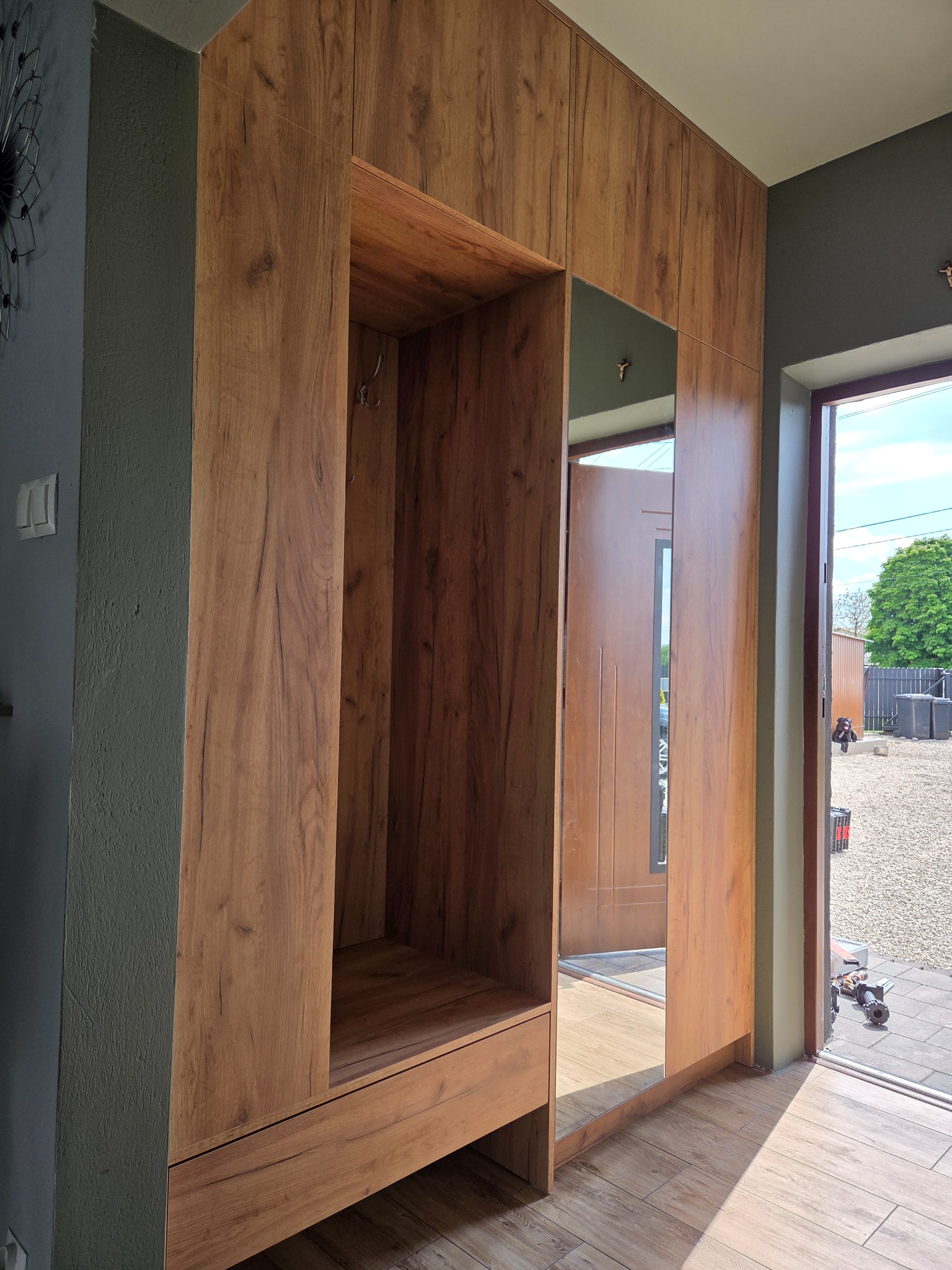 Funkcjonalna szafa wnękowa z lustrem i wieszakami, wykończona w ciepłym odcieniu drewna, idealna do przedpokoju. Widok z perspektywy wejścia do domu.