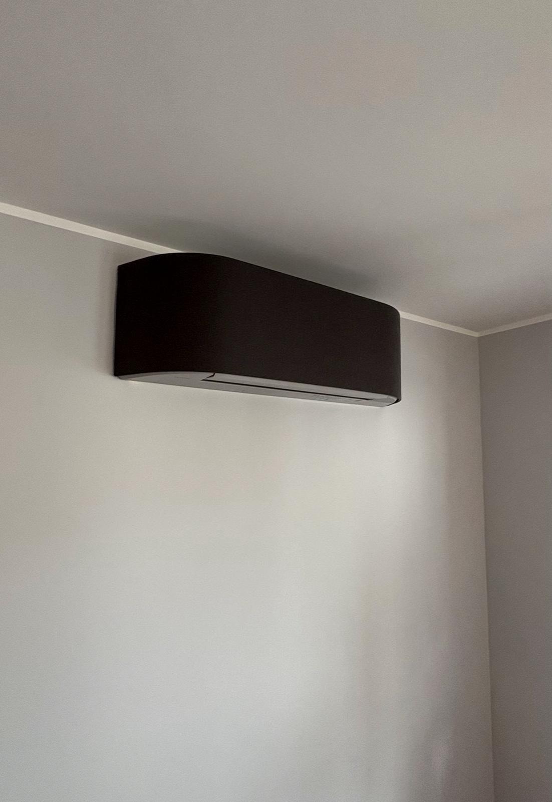 Czarny, nowoczesny klimatyzator ścienny o zaokrąglonych kształtach, zamontowany w rogu jasnego pomieszczenia. Minimalistyczny design urządzenia.