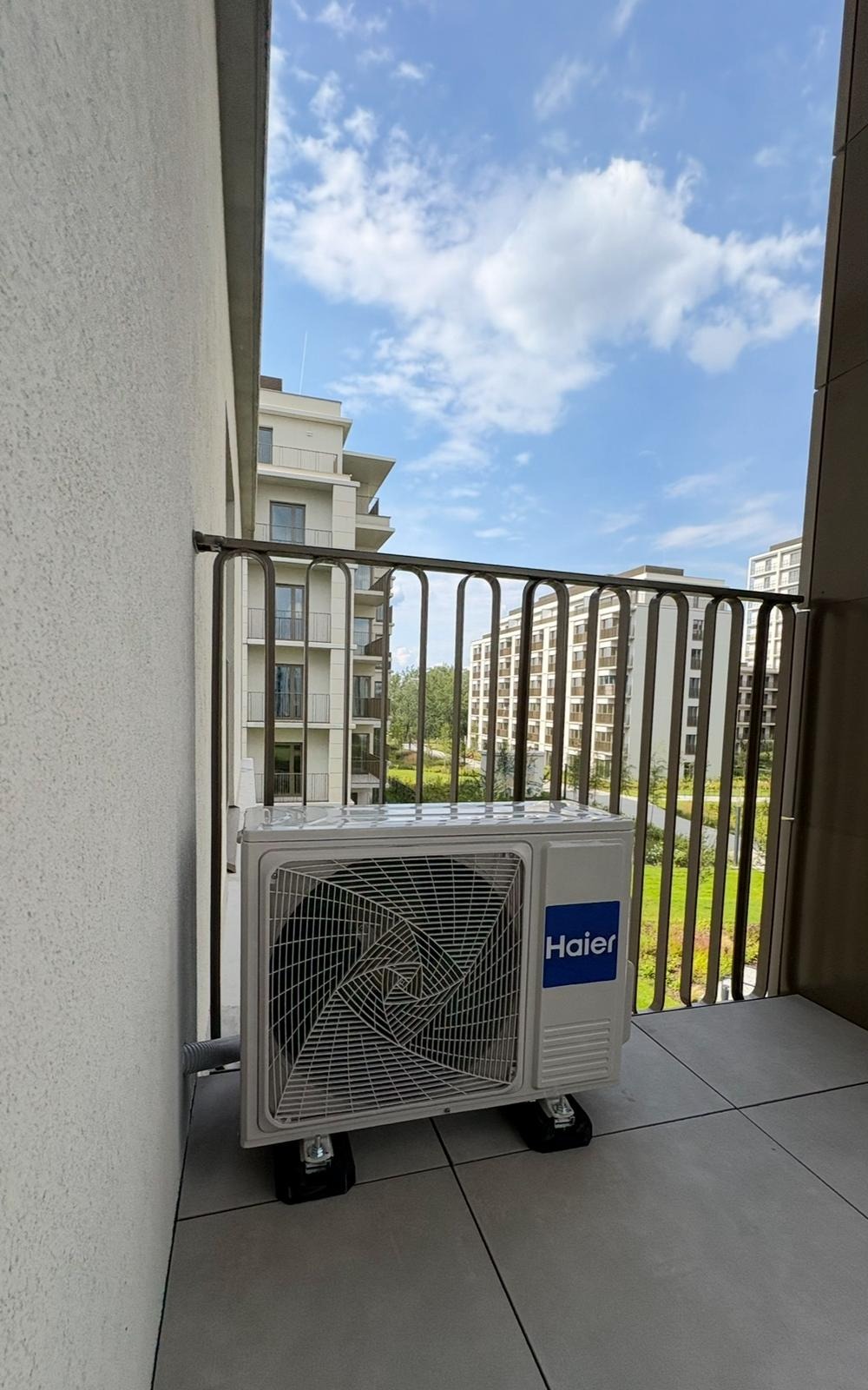 Biała jednostka zewnętrzna klimatyzacji Haier na balkonie nowoczesnego apartamentowca, widok na zieleń i błękitne niebo. Montaż na podkładkach antywibracyjnych.