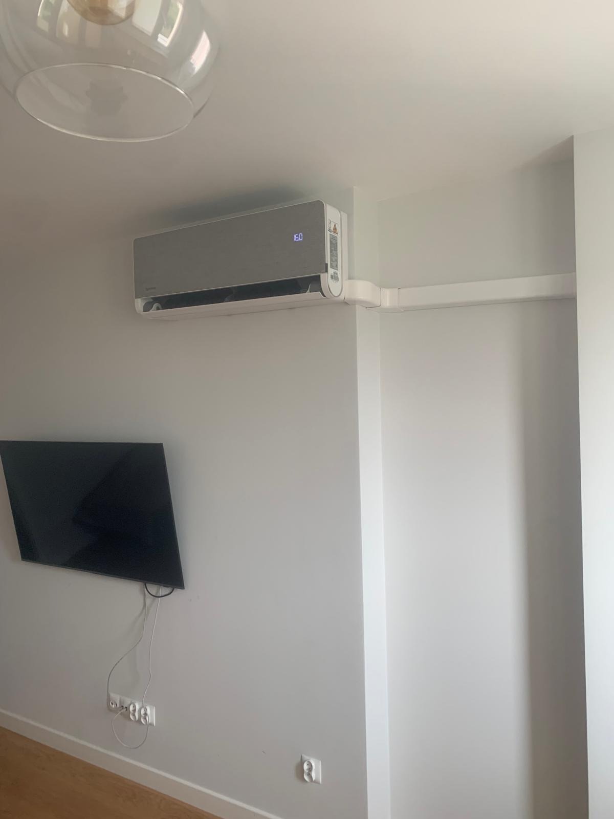 Klimatyzator ścienny w jasnym wnętrzu, z widoczną instalacją elektryczną w białych listwach. Pod spodem telewizor na ścianie. Minimalistyczny design.