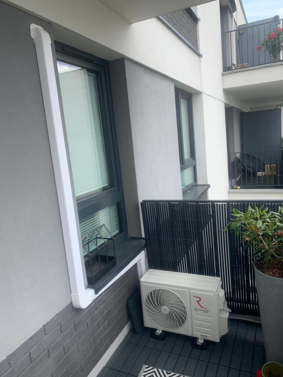 Biała jednostka zewnętrzna klimatyzacji Rotenso na balkonie nowoczesnego budynku, z widoczną instalacją odprowadzającą skropliny wzdłuż okna.