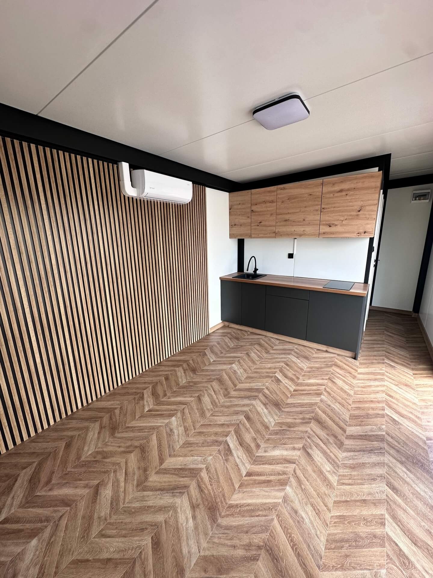Nowoczesne wnętrze z klimatyzacją: drewniana ściana z lamelami, parkiet w jodełkę, zabudowa kuchenna z czarnymi szafkami i drewnianymi górnymi szafkami.