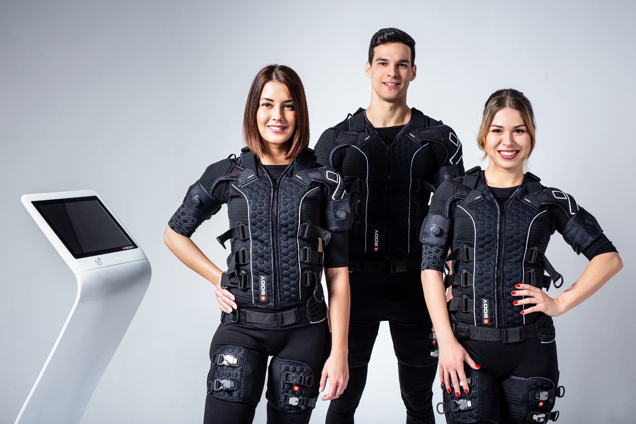 Trzy osoby ubrane w kombinezony EMS, uśmiechnięte, stoją obok białego urządzenia EMS. Widoczne detale kombinezonów i urządzenia.