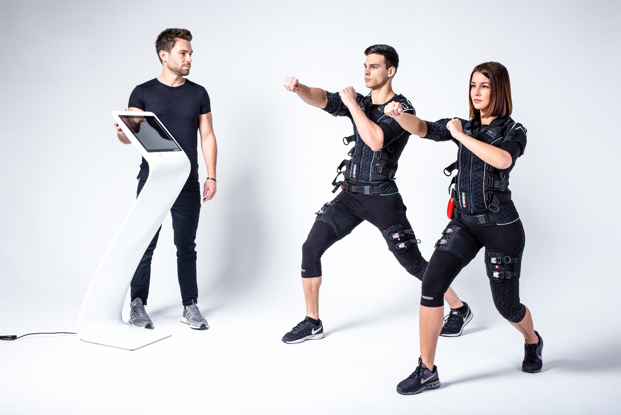 Osoby w strojach EMS ćwiczą pod okiem trenera z tabletem na tle białego studia. Dynamiczna poza, nowoczesny sprzęt. Trening EMS.