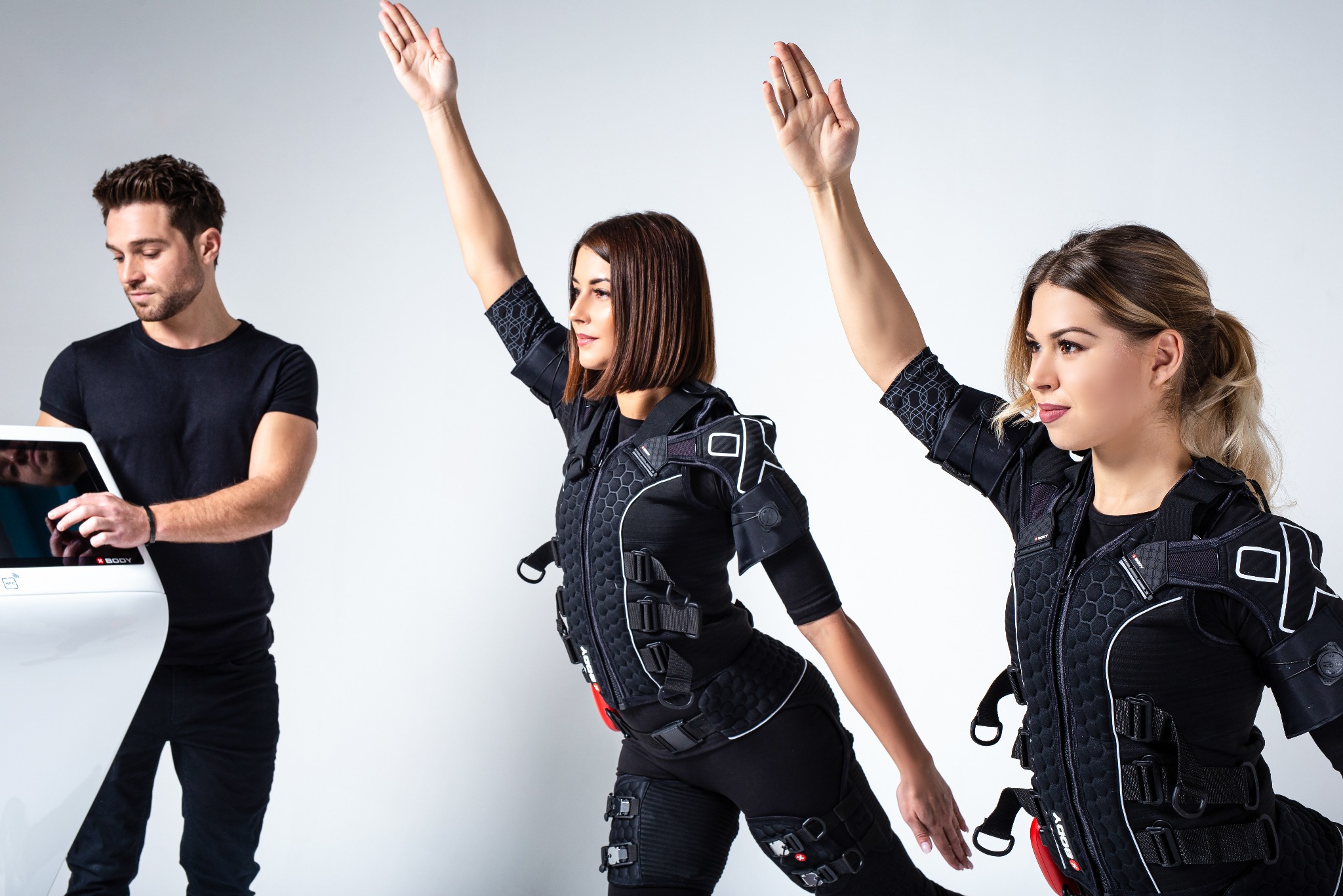 Trening EMS w Warszawie: dwie kobiety w kombinezonach i mężczyzna obsługujący urządzenie. Dynamiczne ujęcie, białe tło, nowoczesny sprzęt. Intensywny trening.