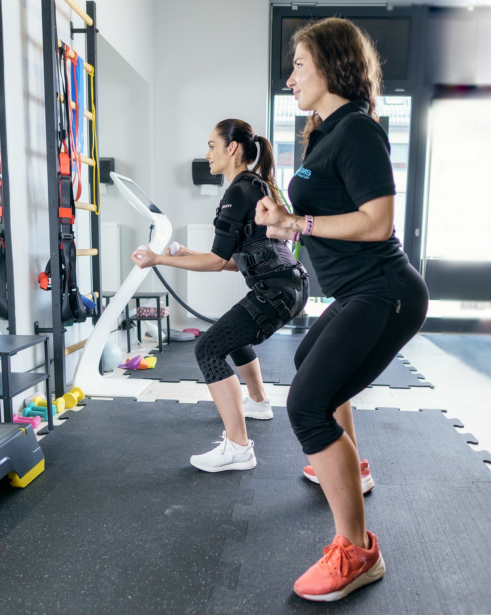 Trening EMS: kobieta w specjalnym stroju ćwiczy przysiady pod okiem trenerki w sportowym stroju. Sprzęt do elektrostymulacji widoczny w tle, drabinki gimnastyczne obok.