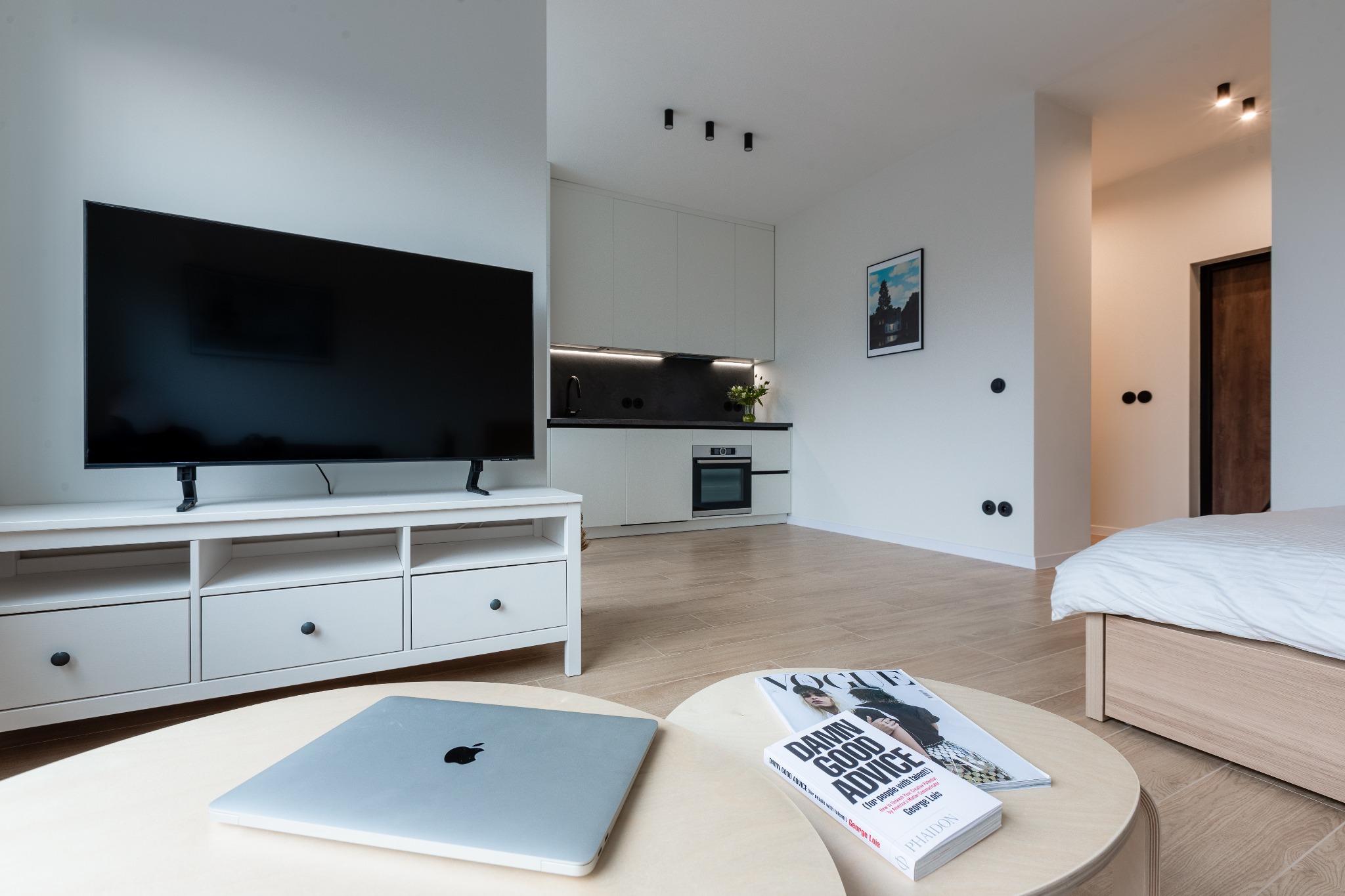 Jasne, nowocześnie urządzone wnętrze mieszkania z aneksem kuchennym, telewizorem i sypialnią w tle. Na okrągłym stoliku leżą laptop i magazyn modowy.