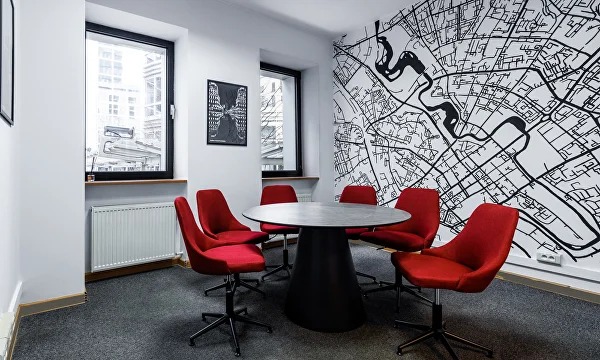 Białe biuro z mapą na ścianie, okrągły stół z czerwonymi krzesłami. Nowoczesny design, czarne ramy okien, szara wykładzina podłogowa. Minimalistyczny wystrój.