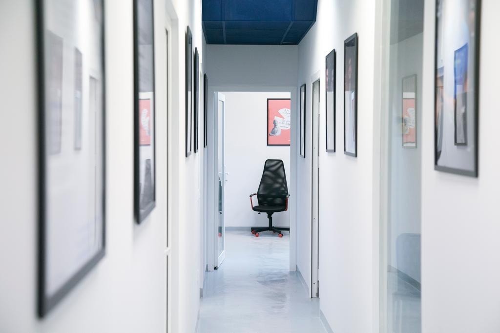 Biały korytarz biurowy z ciemnoniebieskim sufitem, ozdobiony ramkami na ścianach, z krzesłem biurowym w tle, widziany z perspektywy ciągu komunikacyjnego.
