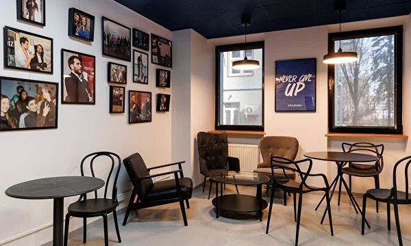 Nowoczesne wnętrze kawiarni z ciemnym sufitem, czarnymi meblami i galerią zdjęć na ścianie, oświetlone lampami wiszącymi. Widok na zewnątrz przez okna.