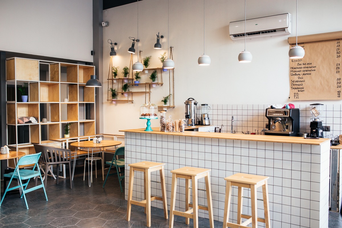 Kawiarnia z drewnianym regałem, stolikami i krzesłami w odcieniach błękitu. Kontuar wyłożony białymi płytkami, na ścianie menu i wiszące lampy.