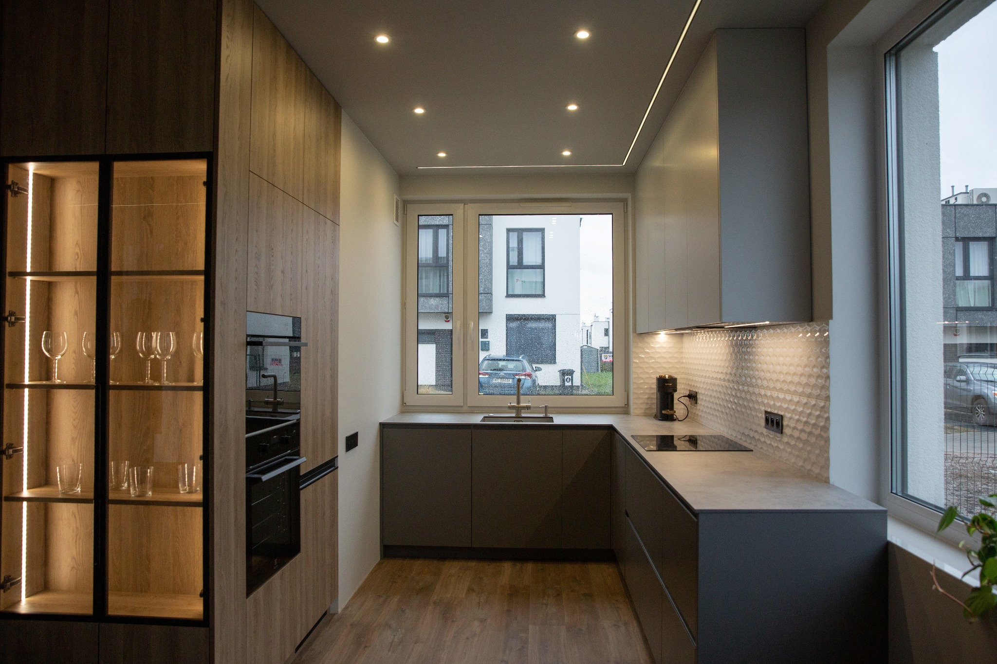 Nowoczesna kuchnia w szarościach z drewnianymi akcentami. Podświetlana witryna z kieliszkami, okno z widokiem na budynek. Minimalistyczny design, oświetlenie LED.