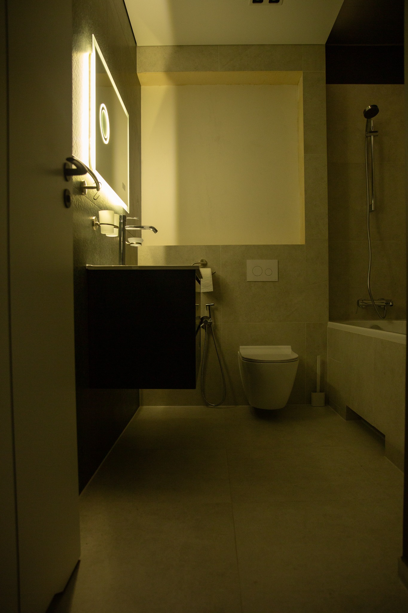 Nowoczesna łazienka w stonowanych barwach z podświetlanym lustrem, wiszącą toaletą i wanną. Minimalistyczny design z elementami szarości i bieli.