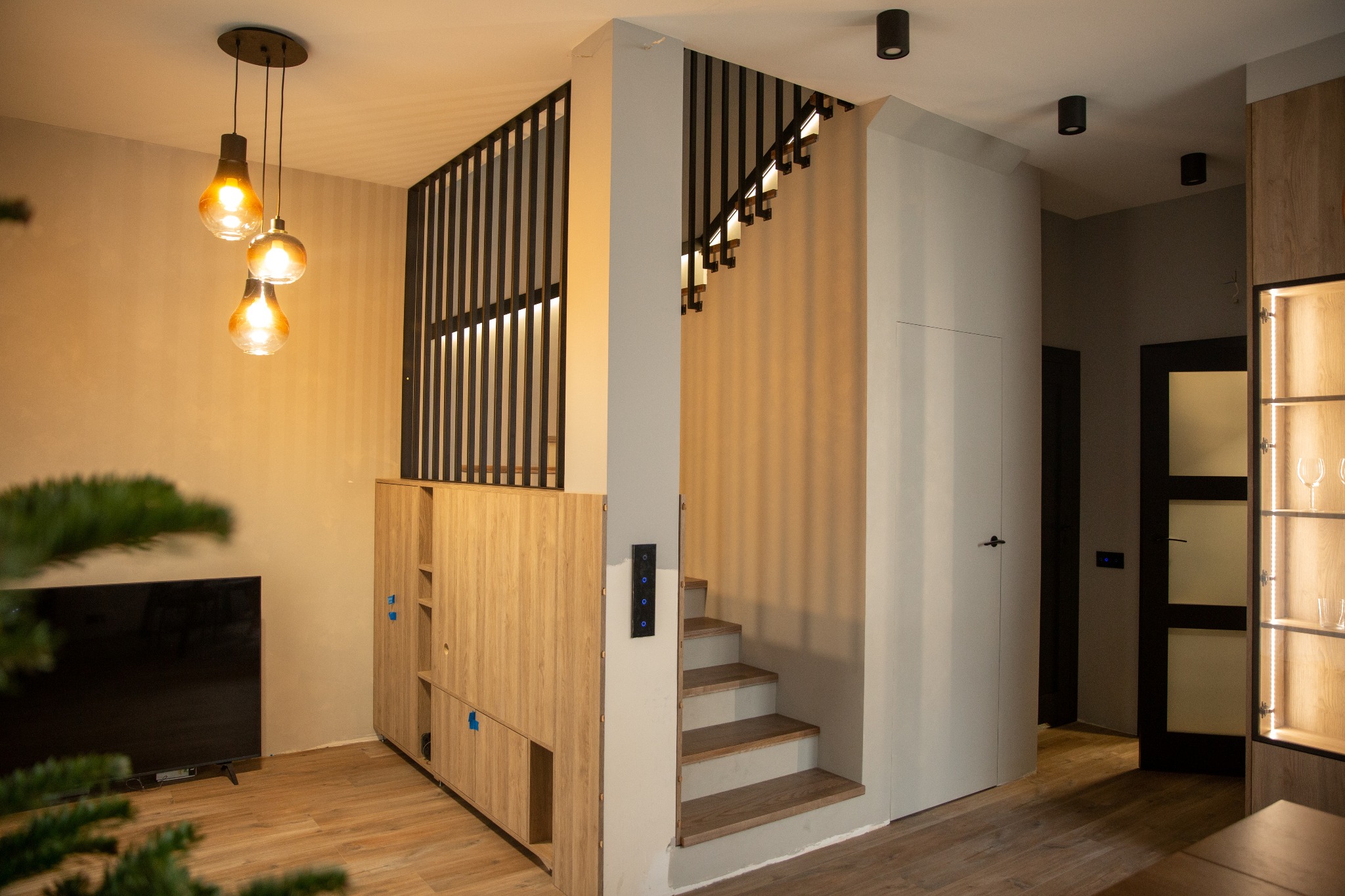 Nowoczesne wnętrze: drewniane schody z czarną balustradą, szafki z oświetleniem i designerskie lampy wiszące w ciepłej tonacji, minimalistyczne wykończenie.