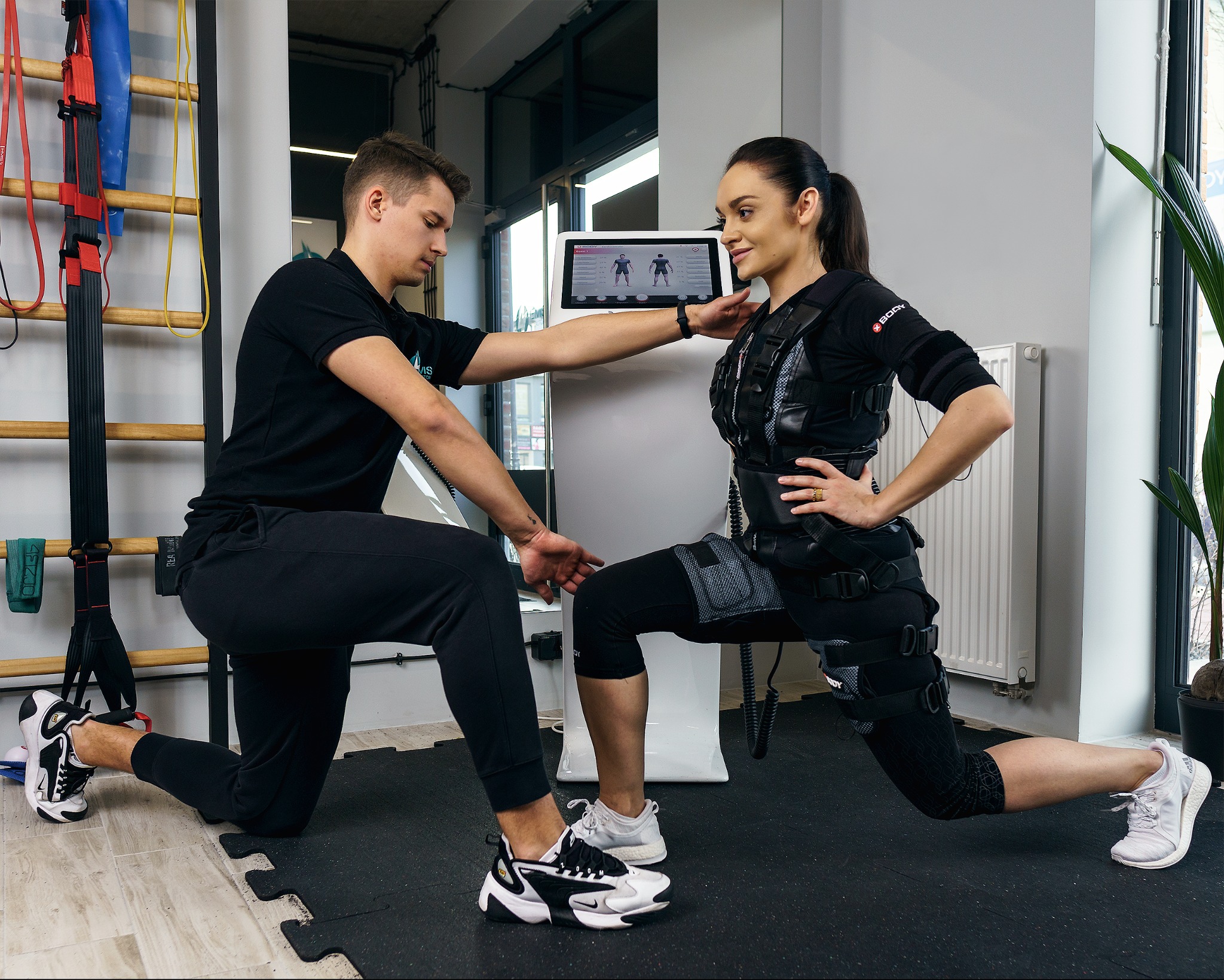 Kobieta w stroju EMS ćwiczy pod okiem trenera. Sprzęt do elektrostymulacji, drabinka gimnastyczna w tle. Trening personalny z wykorzystaniem technologii EMS.