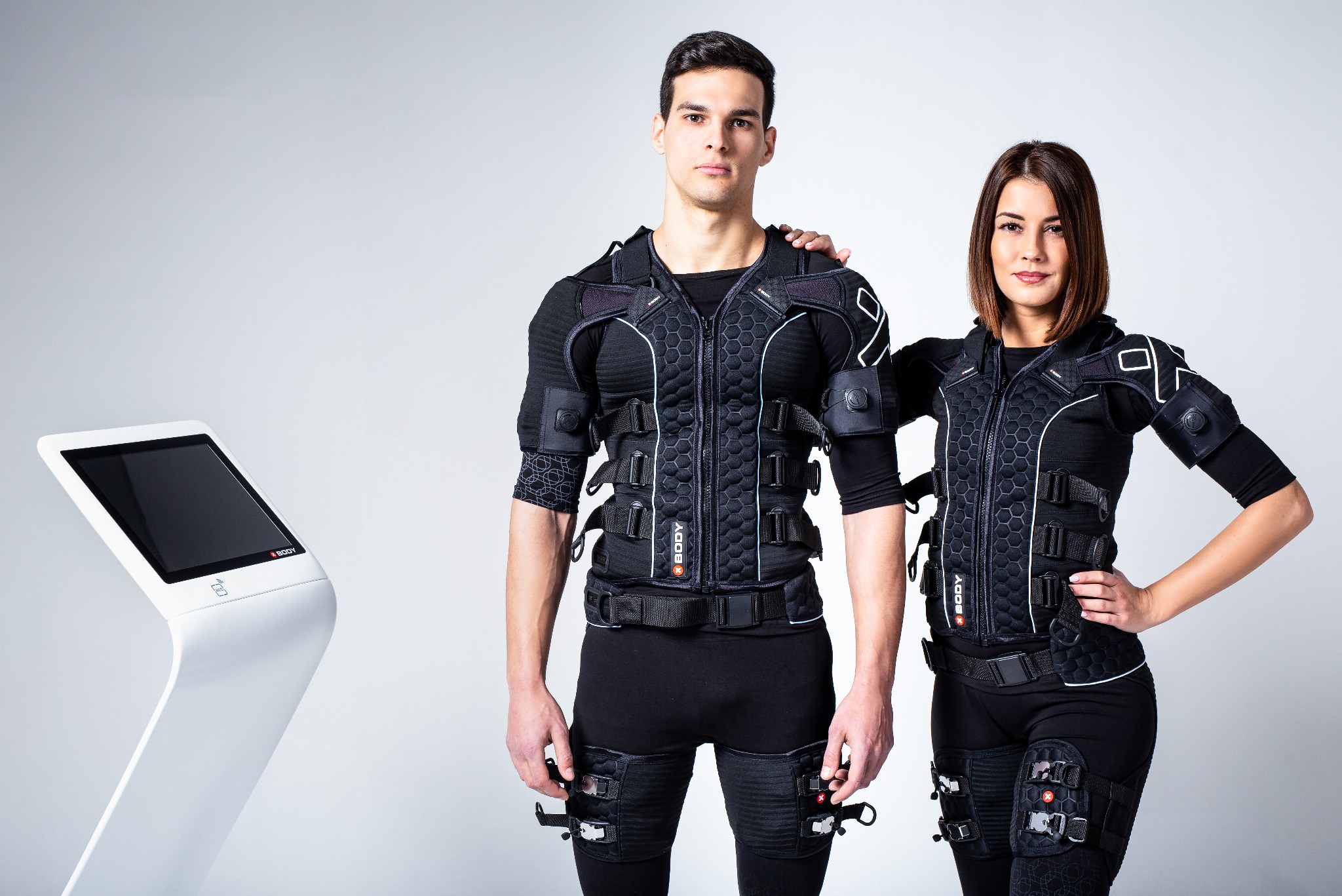 Dwoje uśmiechniętych ludzi w strojach do elektrostymulacji mięśni (EMS) pozuje obok białego urządzenia sterującego. Specyficzne detale stroju i urządzenia.