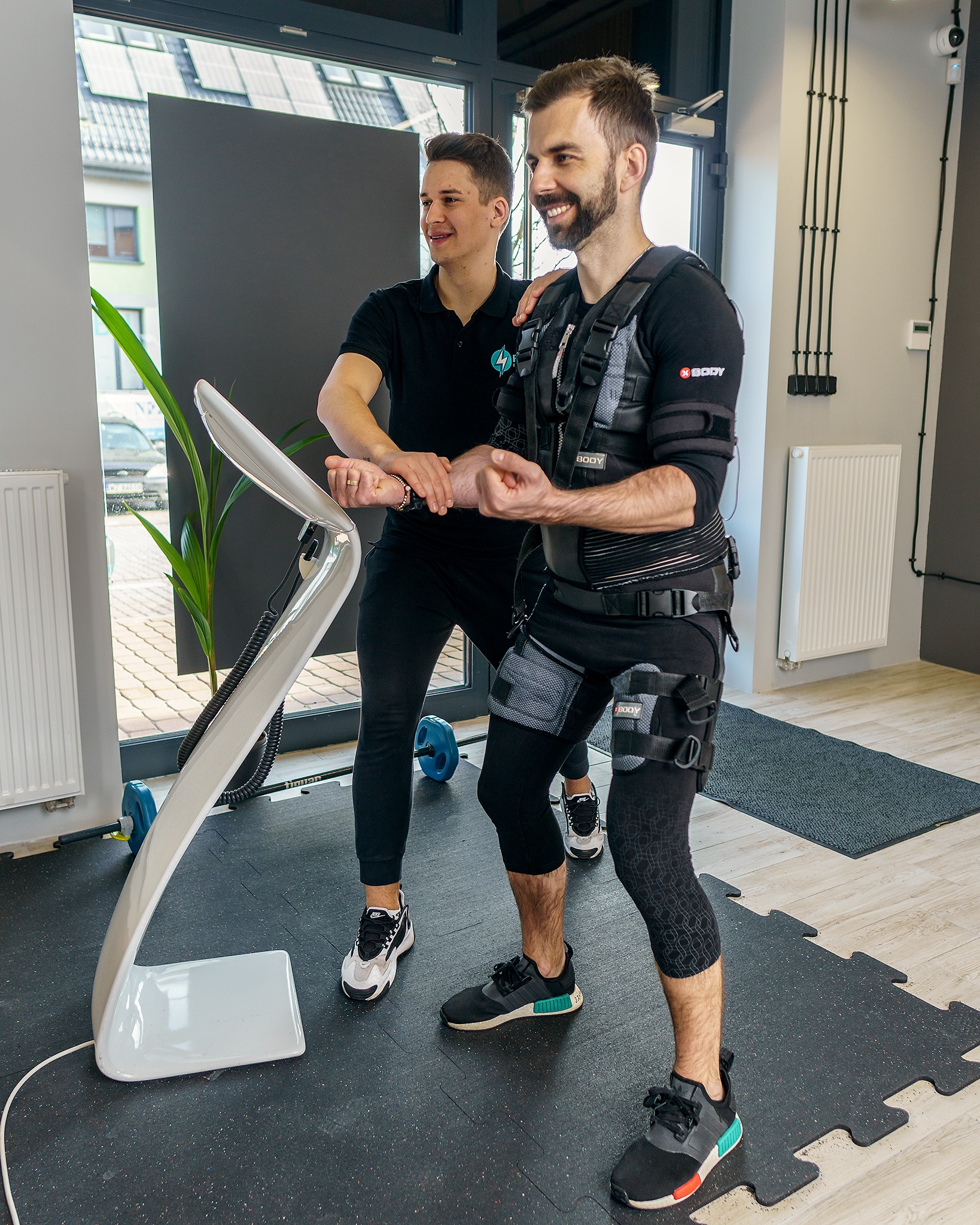 Trening EMS: uśmiechnięty mężczyzna w specjalnym stroju ćwiczy pod okiem trenera. Nowoczesne studio fitness, sprzęt do elektrostymulacji mięśni.
