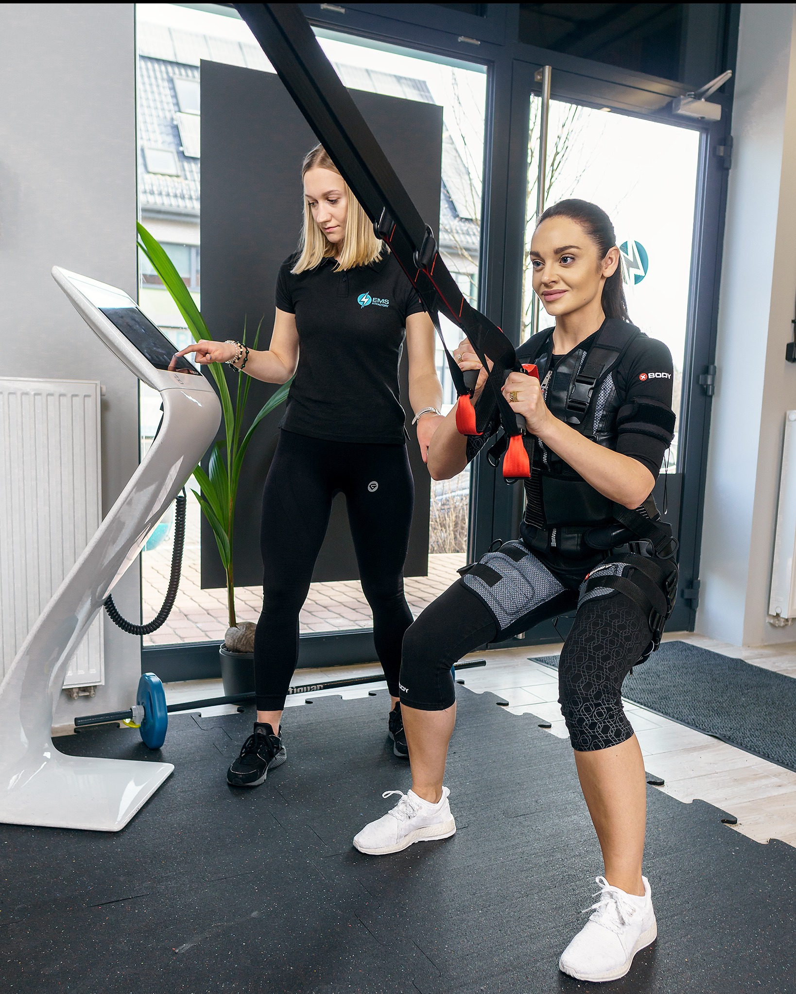 Trening EMS: kobieta w specjalnym kombinezonie ćwiczy pod okiem trenerki, która obsługuje urządzenie do elektrostymulacji. Nowoczesne studio fitness.