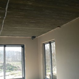 PROBUD ST SPÓŁKA Z OGRANICZONĄ ODPOWIEDZIALNOŚCIĄ - Wnętrze po szpachlowaniu w Sierakowicach. Widoczne gładkie ściany, surowy betonowy sufit z instalacją elektryczną i dwa okna z widokiem na zewnątrz. Jasne, naturalne światło.