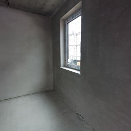 PROBUD ST SPÓŁKA Z OGRANICZONĄ ODPOWIEDZIALNOŚCIĄ - Surowe wnętrze po tynkowaniu maszynowym, widoczne okno z białymi ramami. Szare ściany i podłoga, przygotowane do dalszych prac wykończeniowych.